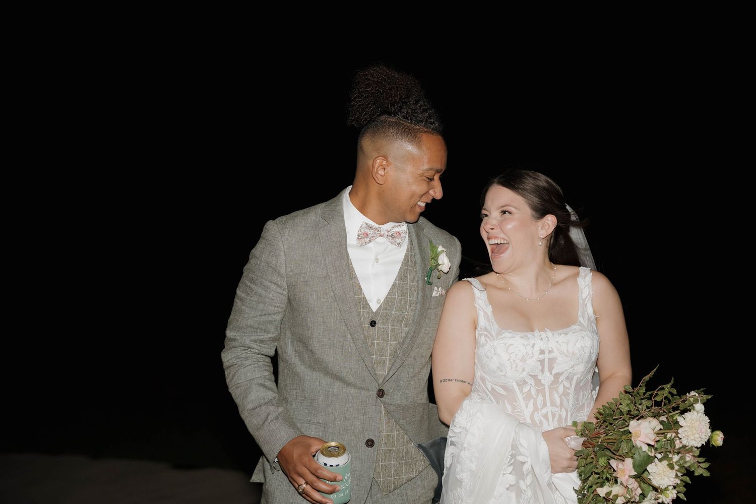 A couple shares a joyful moment in wedding attire with bride wearing white dress and groom in gray vest and bowtie.