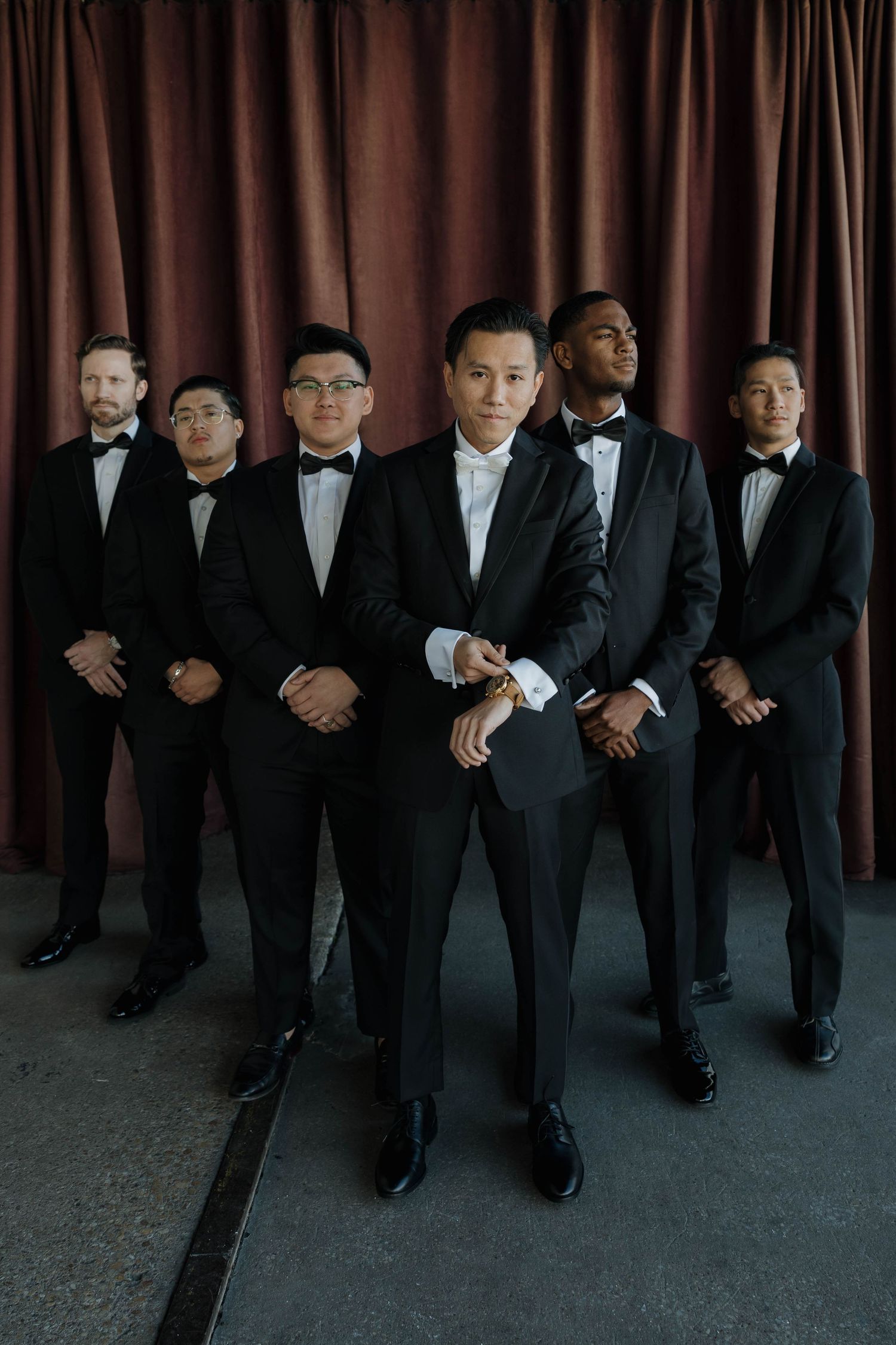 Groomsmen wearing black suits and bowties stand in stylish formation against a burgundy curtain backdrop.