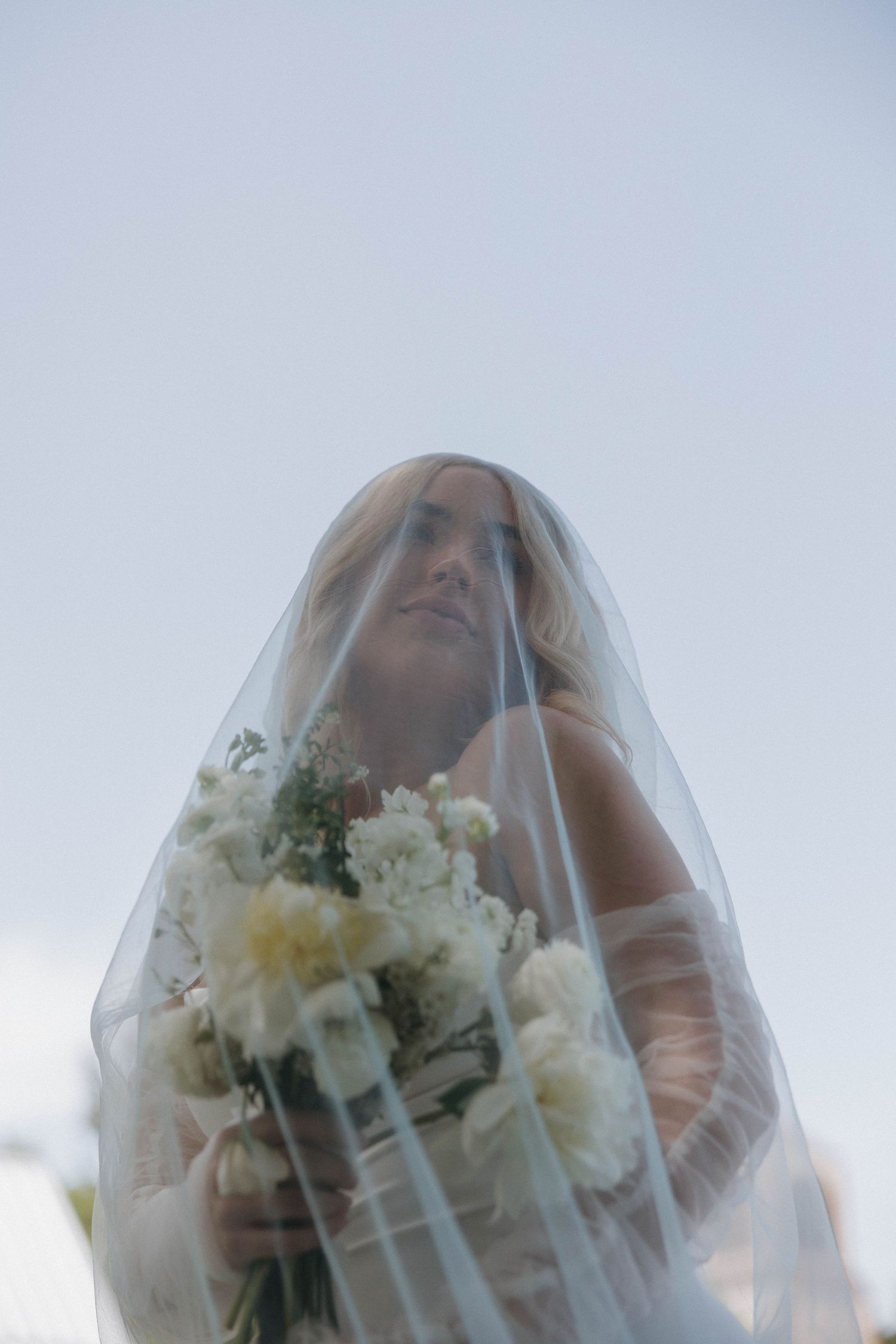 A bride in a traditional white veil holds a romantic bouquet of ivory and white flowers against a soft sky background.