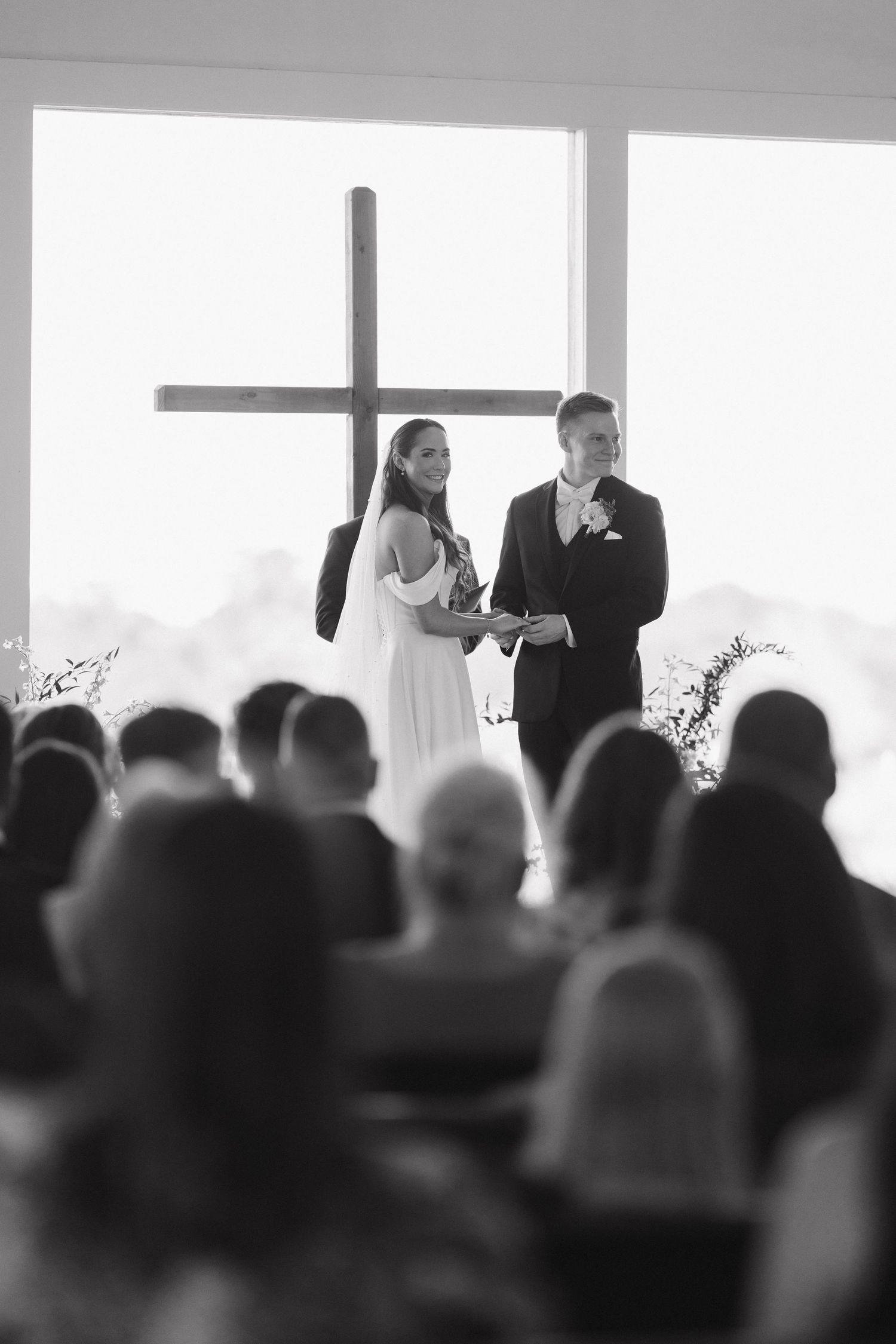 Black and white wedding ceremony photo showing a couple standing at an altar with a large cross in the background and guests seated.