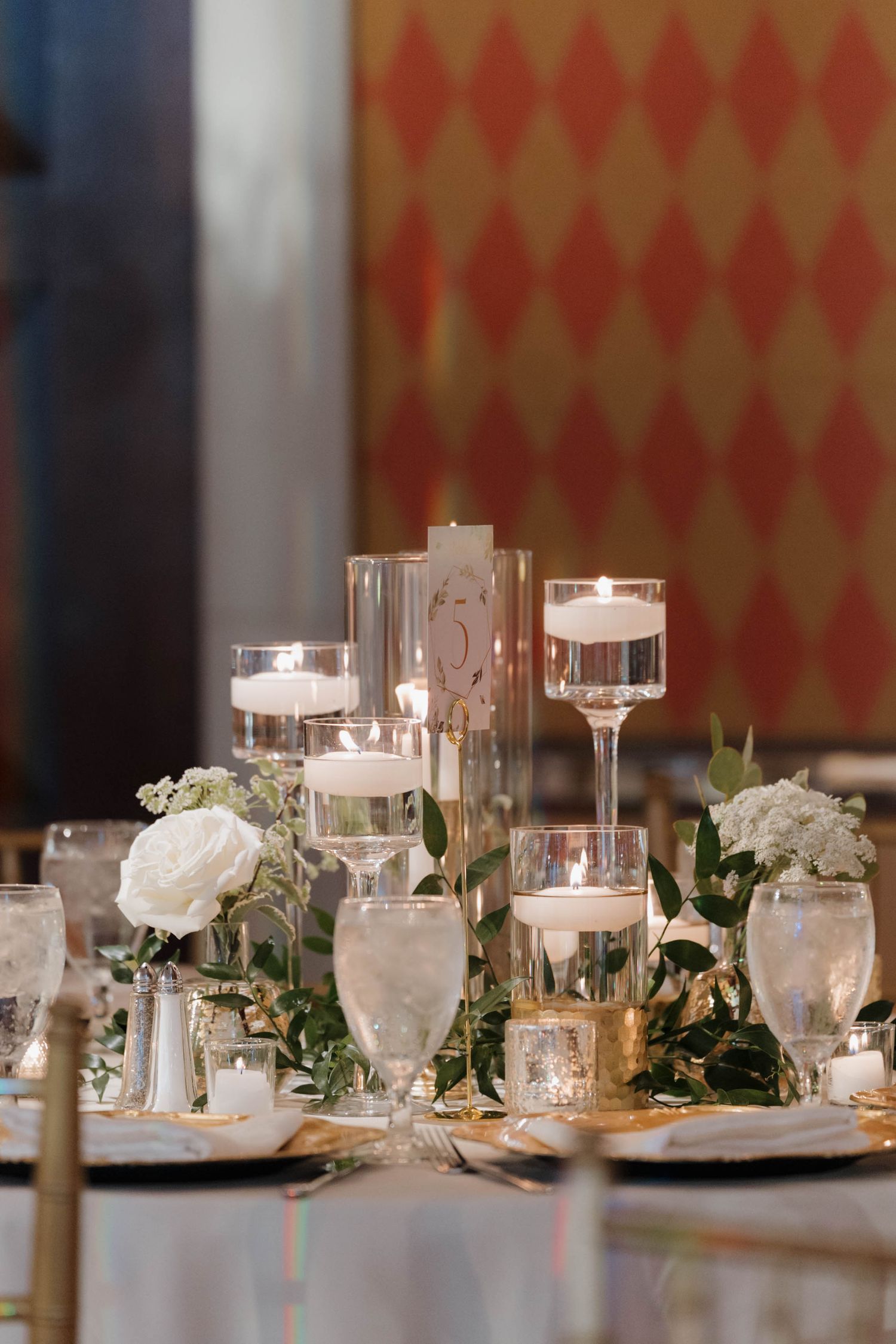 Elegant wedding table setting with floating candles in glass holders, white roses, and greenery against a red patterned wall.
