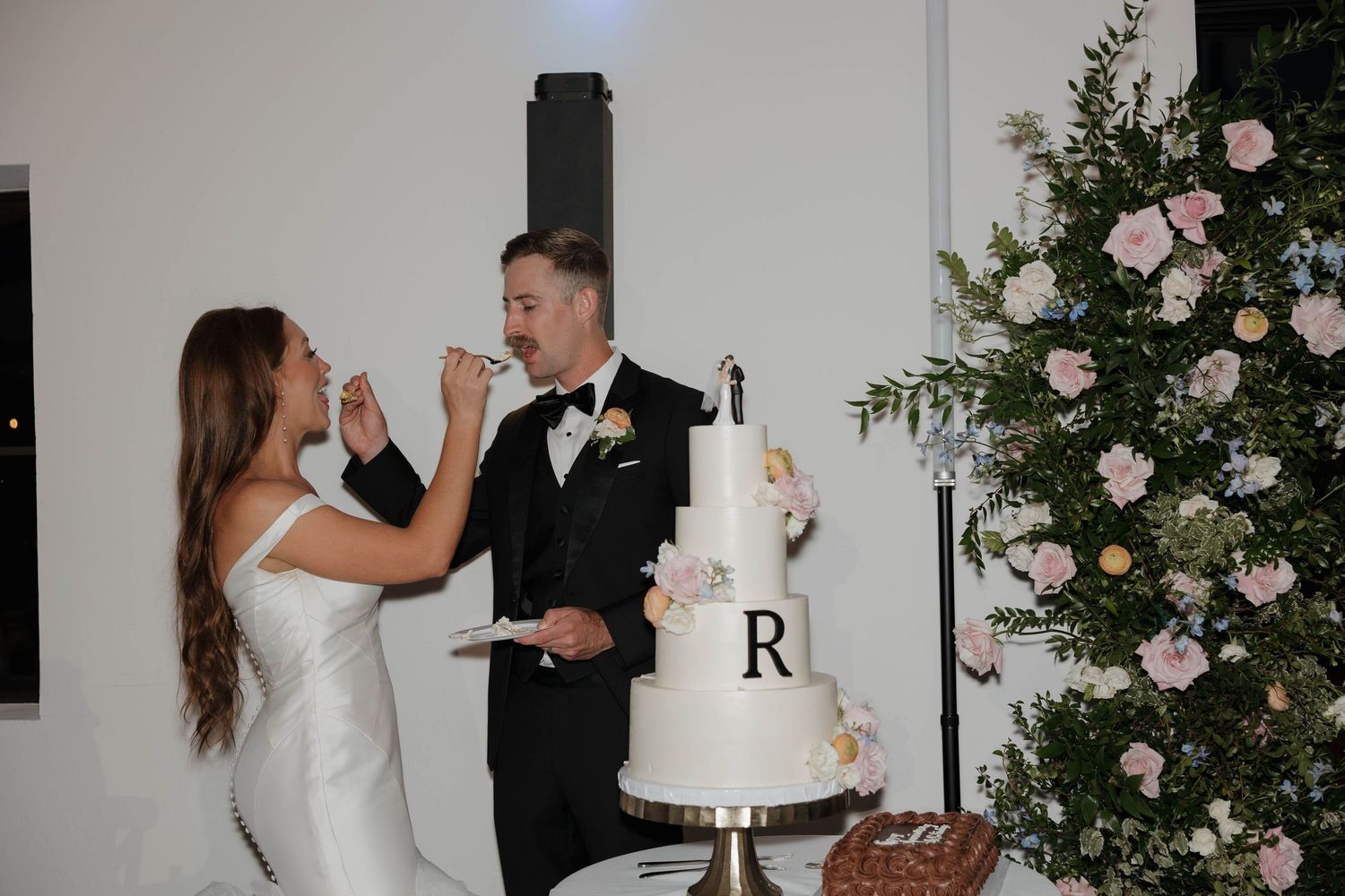 Newlyweds playfully feed each other wedding cake beside a white tiered cake and pink rose floral arrangement.