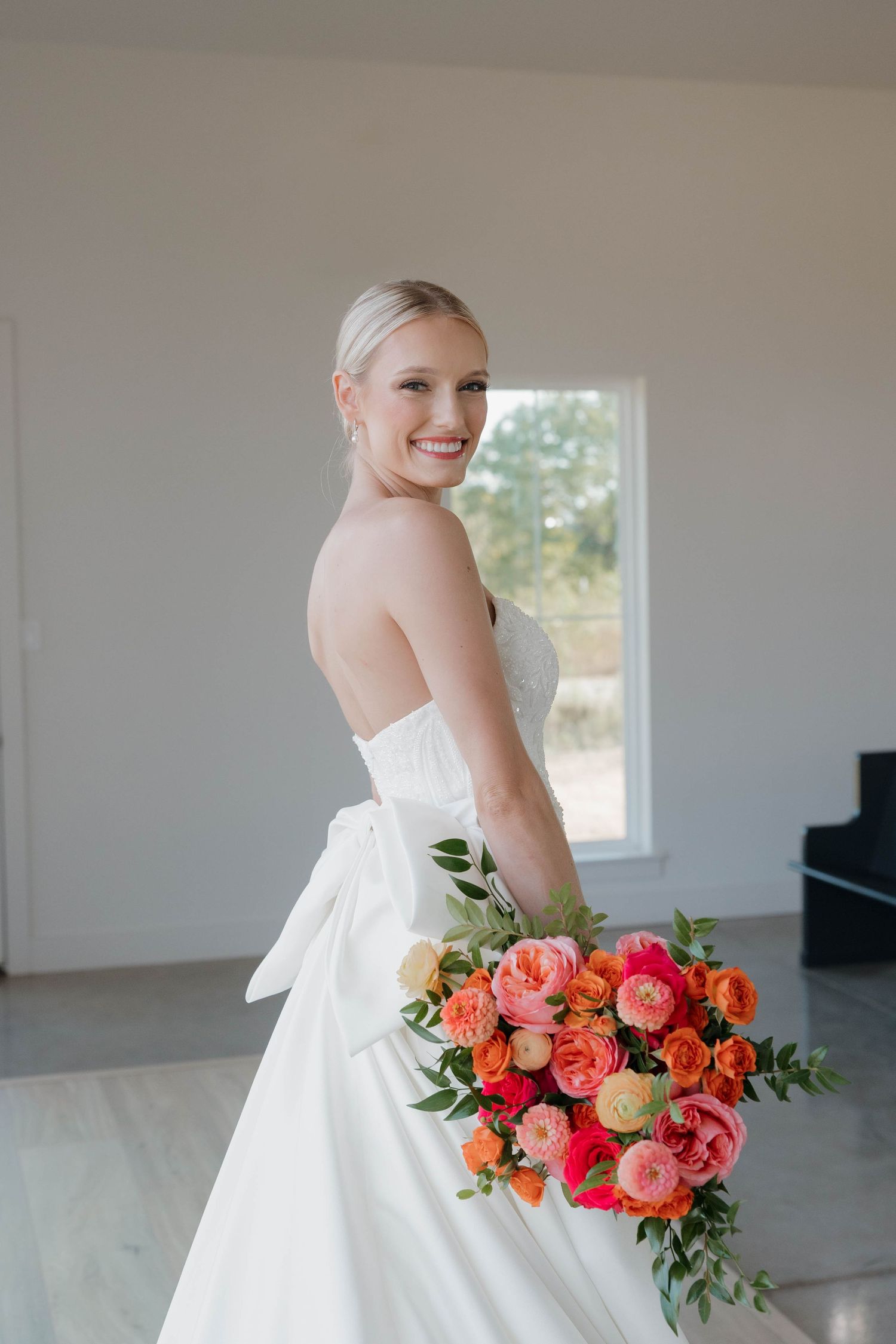 Bride in white wedding gown holds vibrant coral and red bouquet against bright interior.