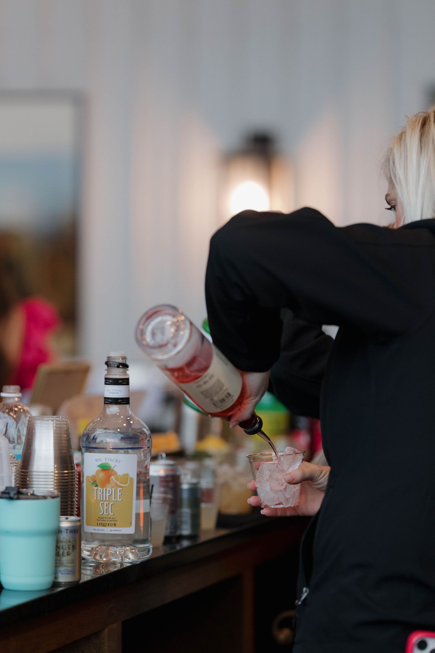 Bartender in black pouring a cocktail at a bar counter with various bottles and mixers displayed.