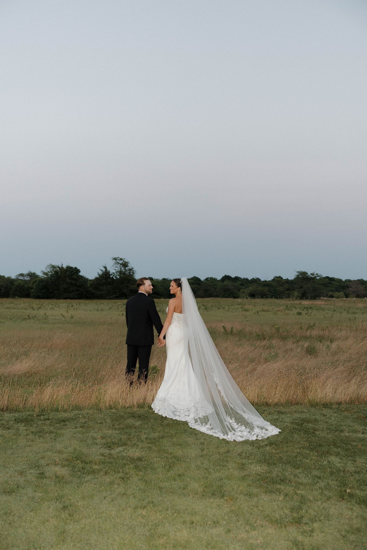 A bride and groom share an intimate moment in a golden grass field during sunset with a long flowing veil.