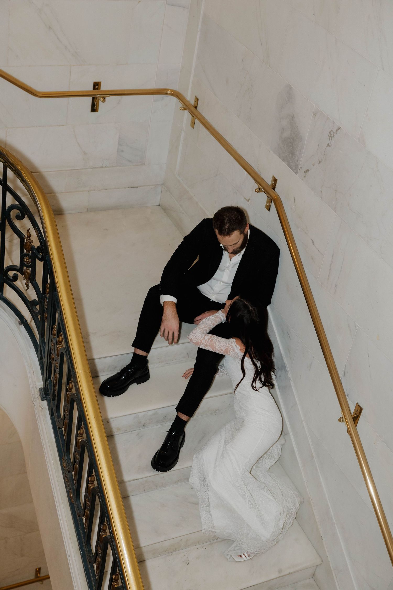 A wedding couple rests dramatically on marble stairs with gold railings.