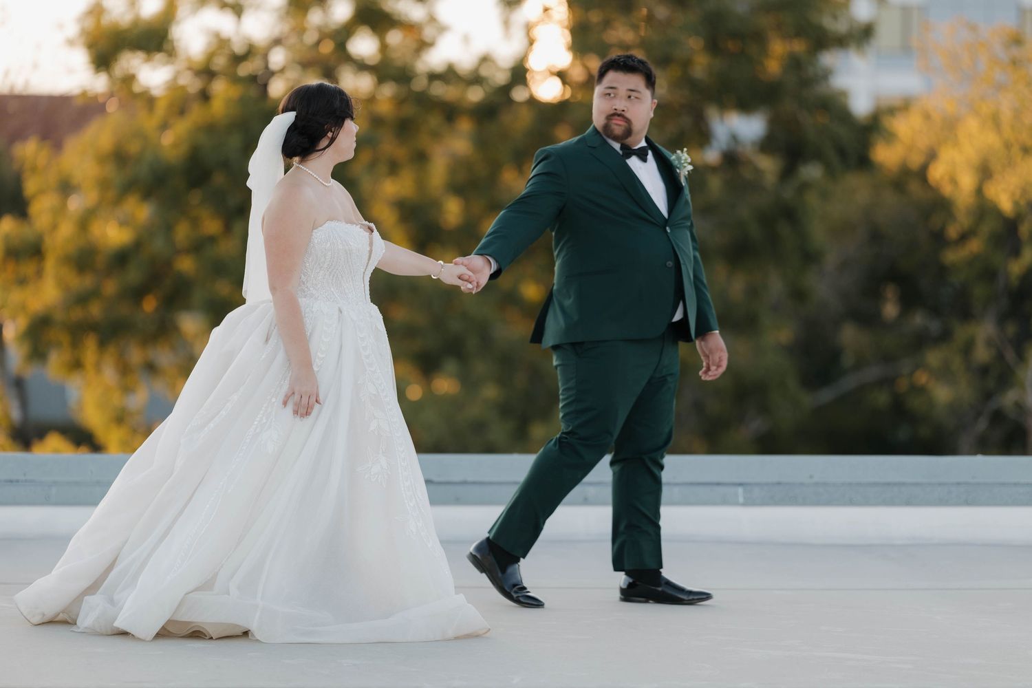 A couple in wedding attire dances together on a light colored floor during golden hour with autumn trees in background.