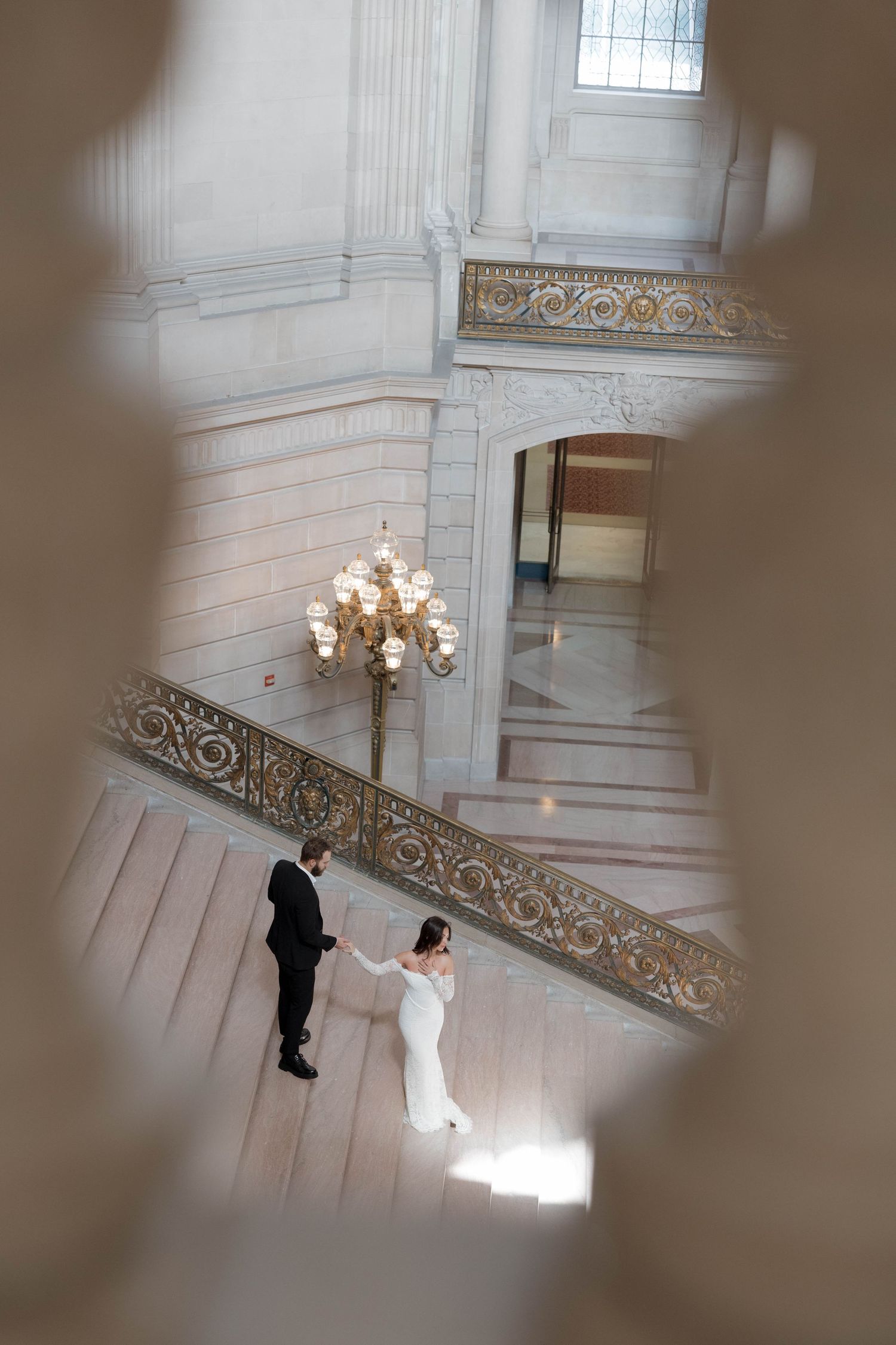 Two people in formal attire stand on elegant marble stairs beneath an ornate chandelier at a grand historic venue.