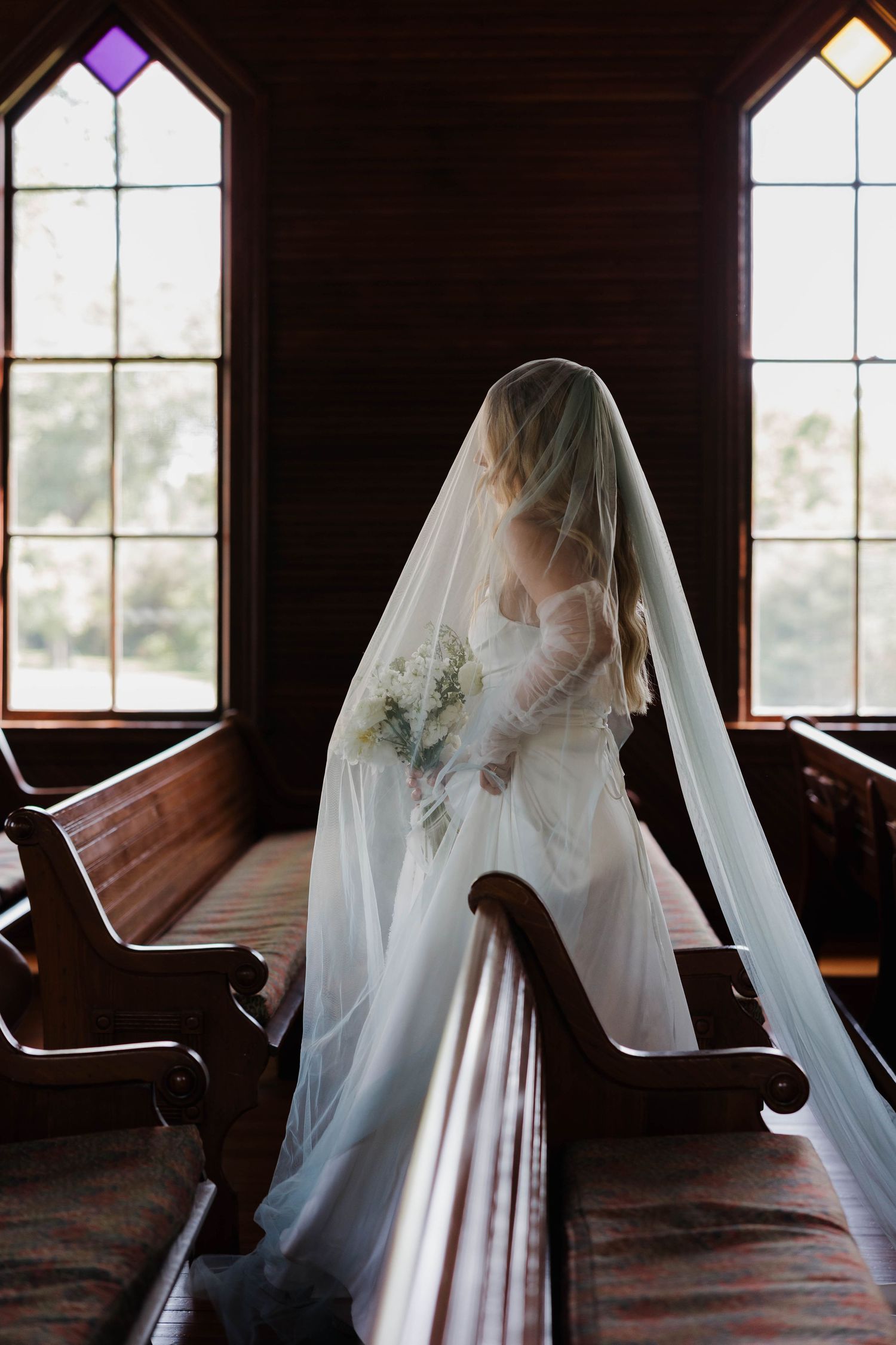 A bride in a white gown and flowing veil stands gracefully between wooden church pews near gothic windows.