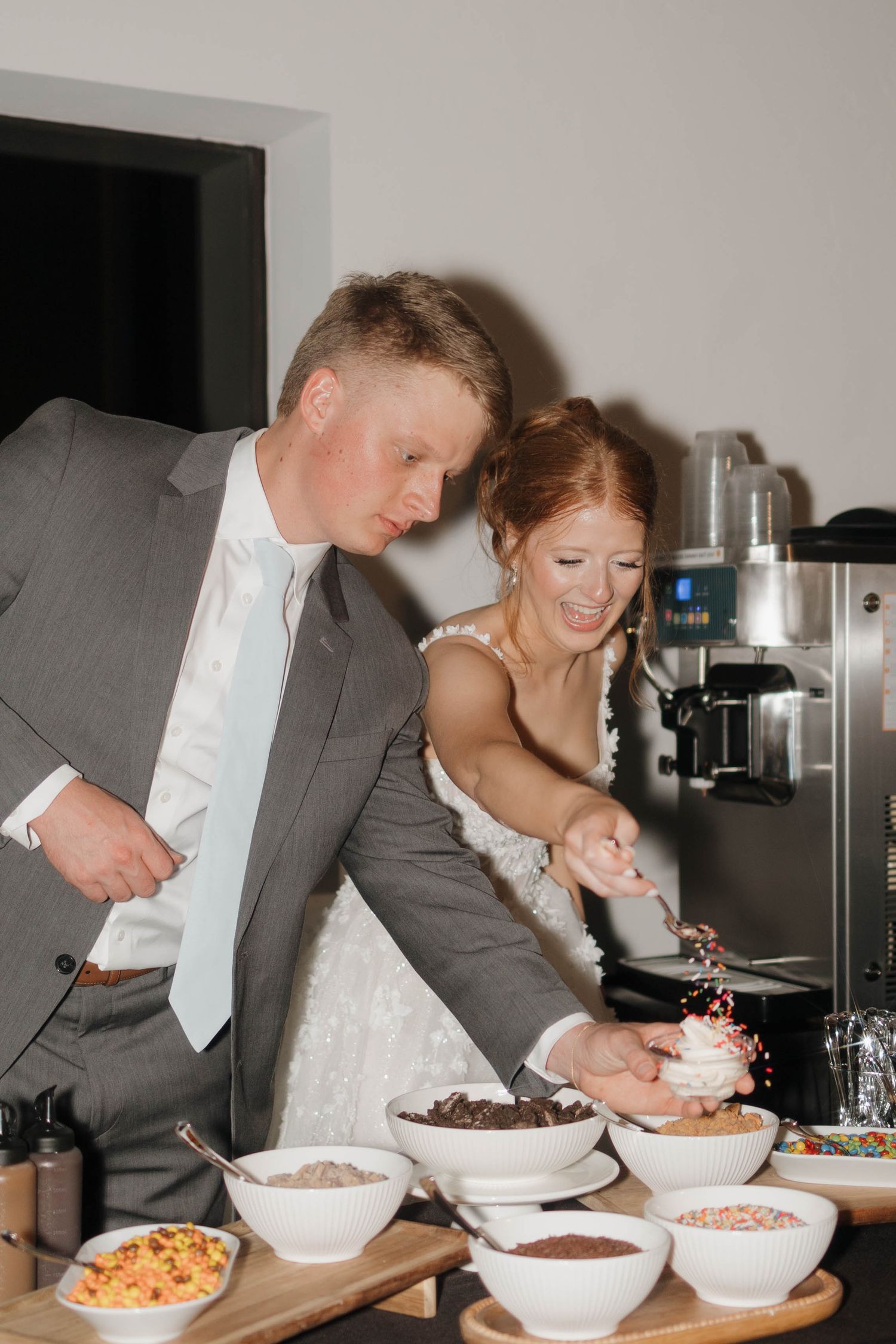 A couple in formal attire joyfully sample various snacks and treats from multiple white serving bowls at a reception.
