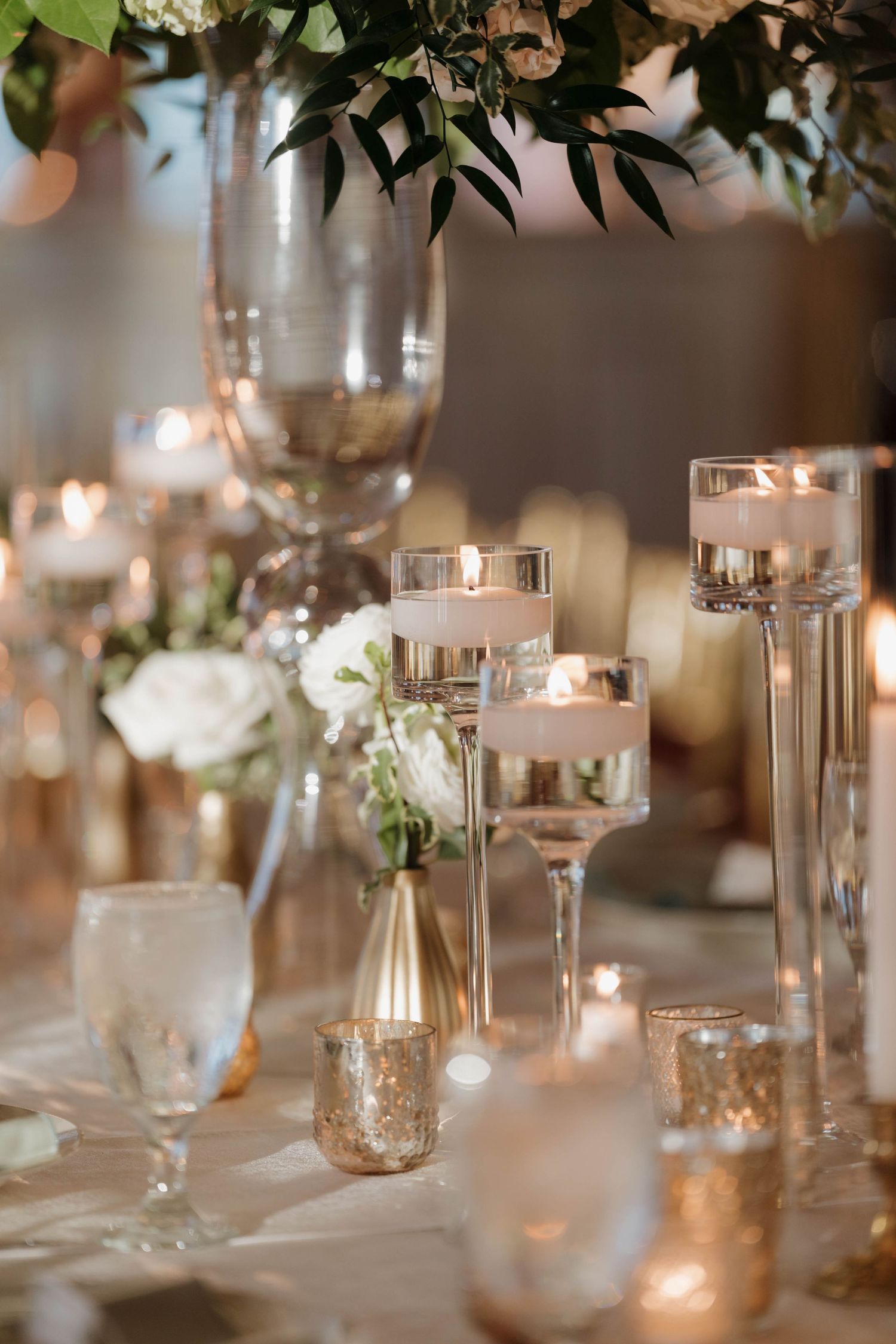 Elegant wedding reception table settings with glowing candles and delicate white floral arrangements in glass vessels.