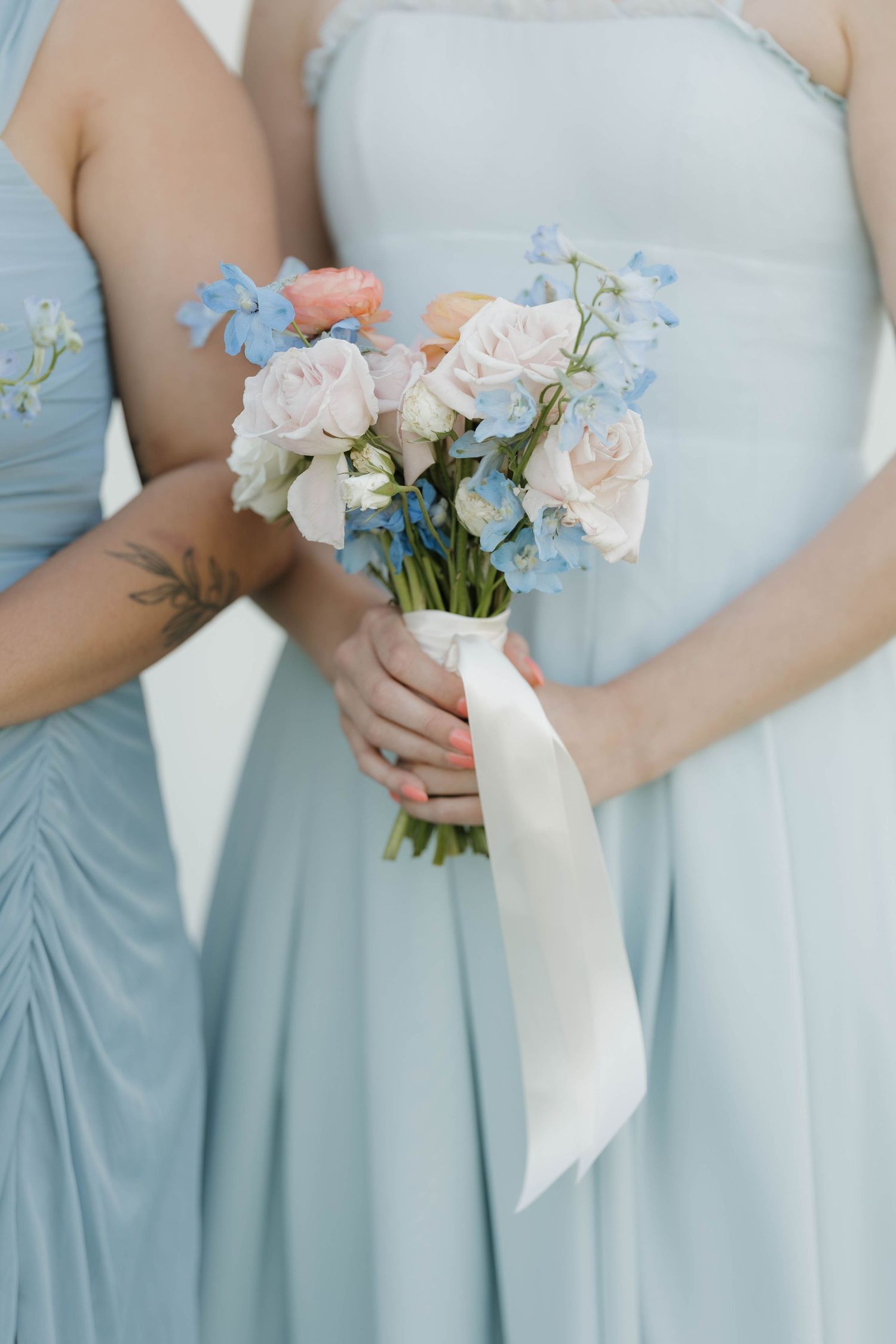 A delicate bouquet of blush roses and blue delphiniums held against powder blue bridesmaid dresses with white ribbon.