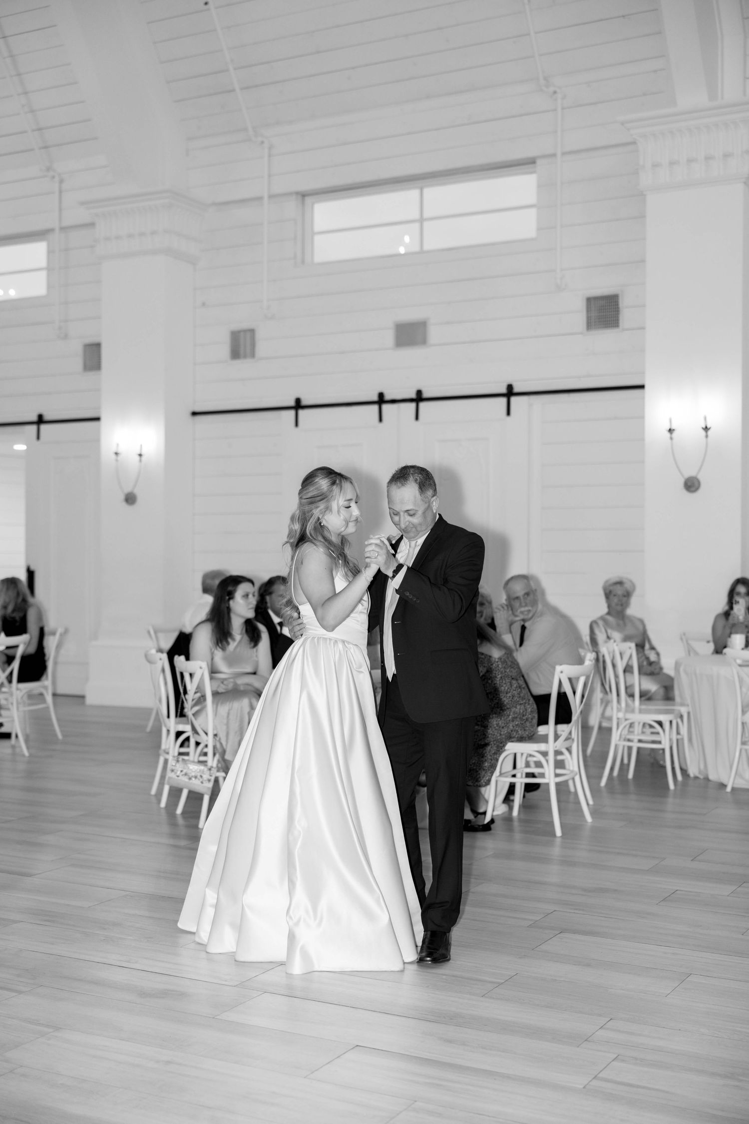 A black and white photo captures a couple sharing their first dance in an elegant white reception venue.