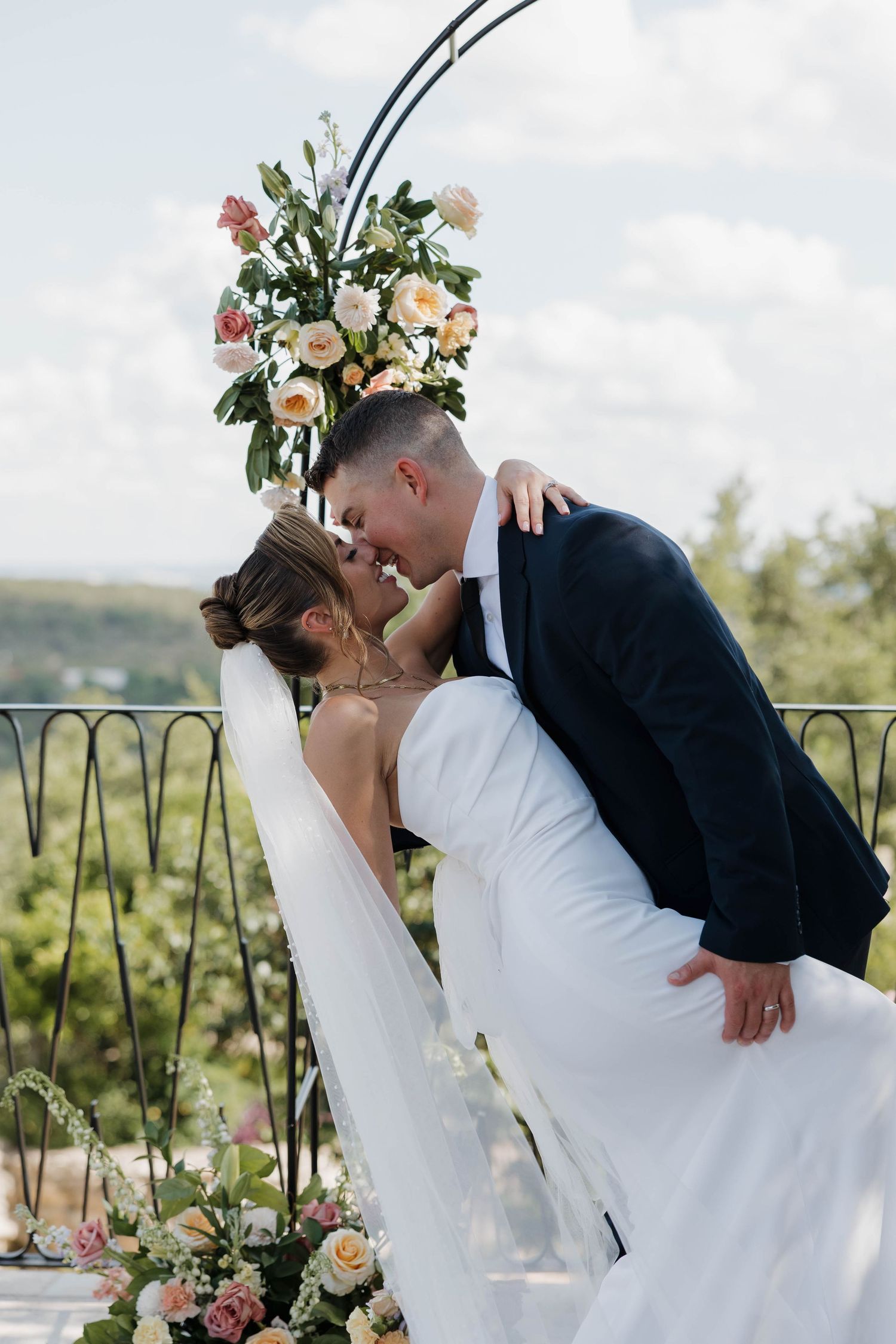 The couple shares a romantic dip kiss with the bride wearing a floral crown and white dress against an outdoor backdrop.