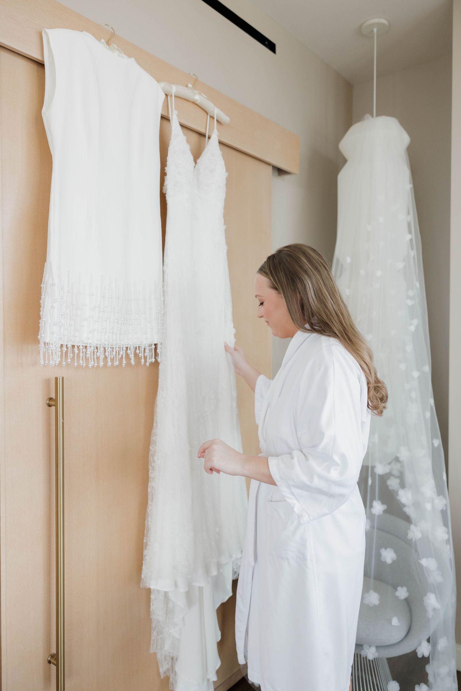 Woman in white robe looking at wedding dresses hanging on wall in bridal boutique shop.