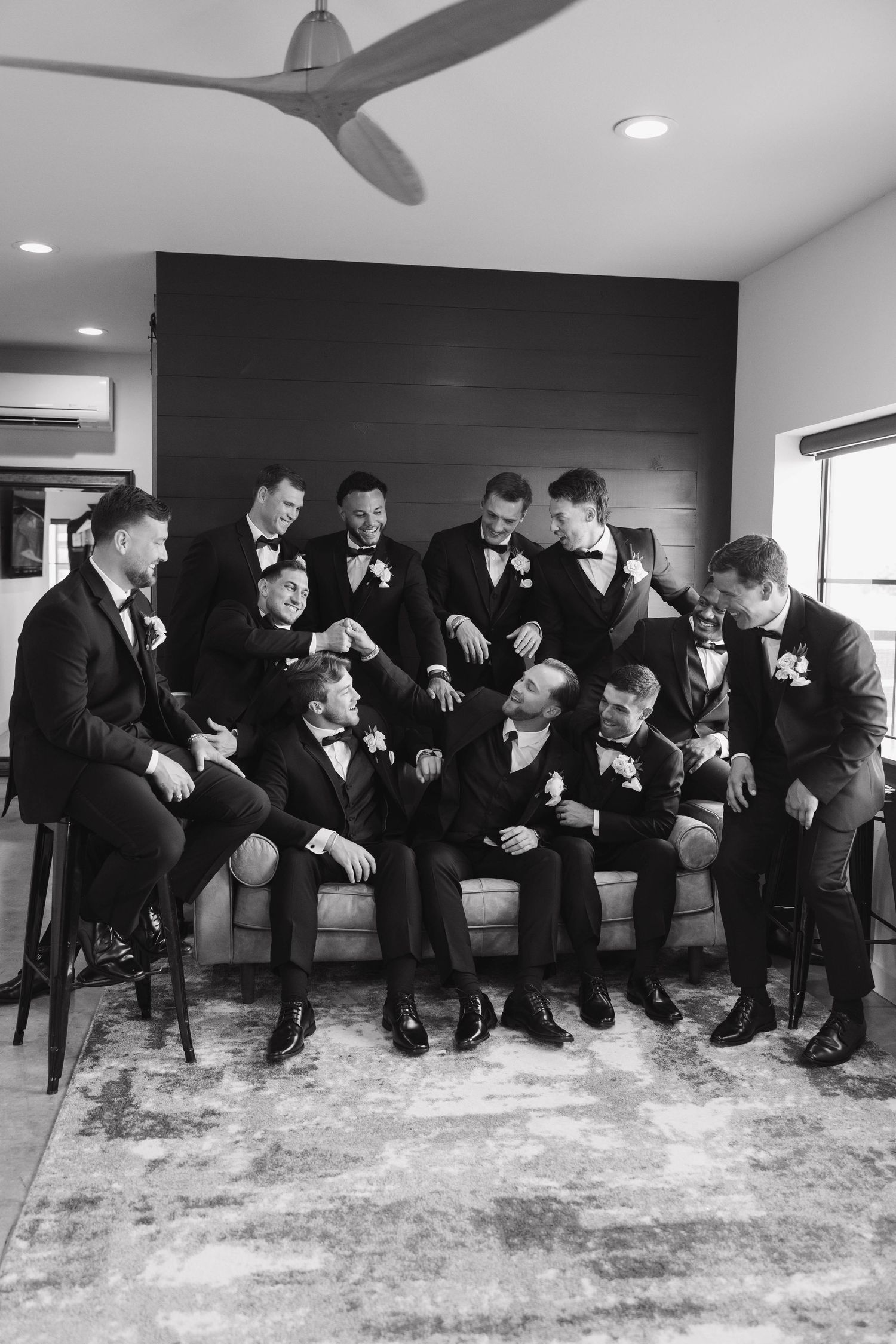 Large group of groomsmen in black tuxedos laughing and bonding together in contemporary room with modern decor.