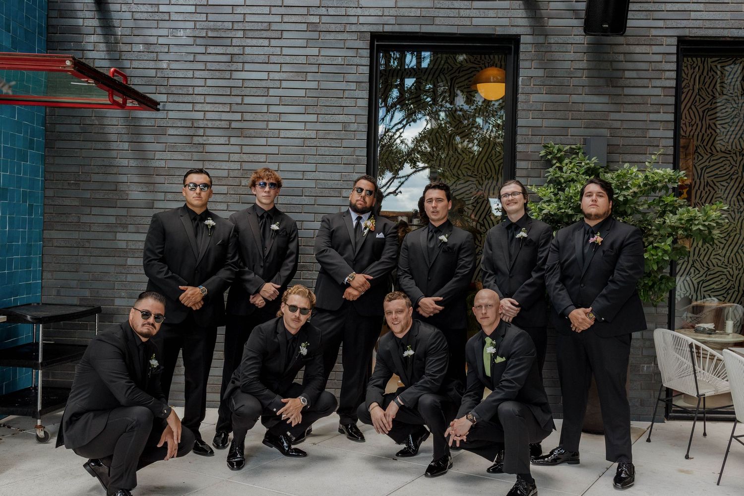 A wedding party dressed in all black formal attire poses together outdoors in front of a brick wall.