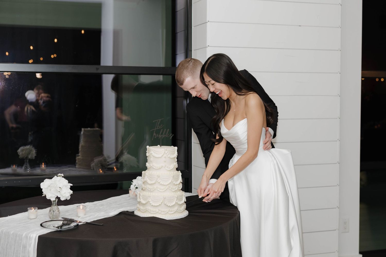 A couple cutting their elegant white wedding cake together at an evening reception celebration.