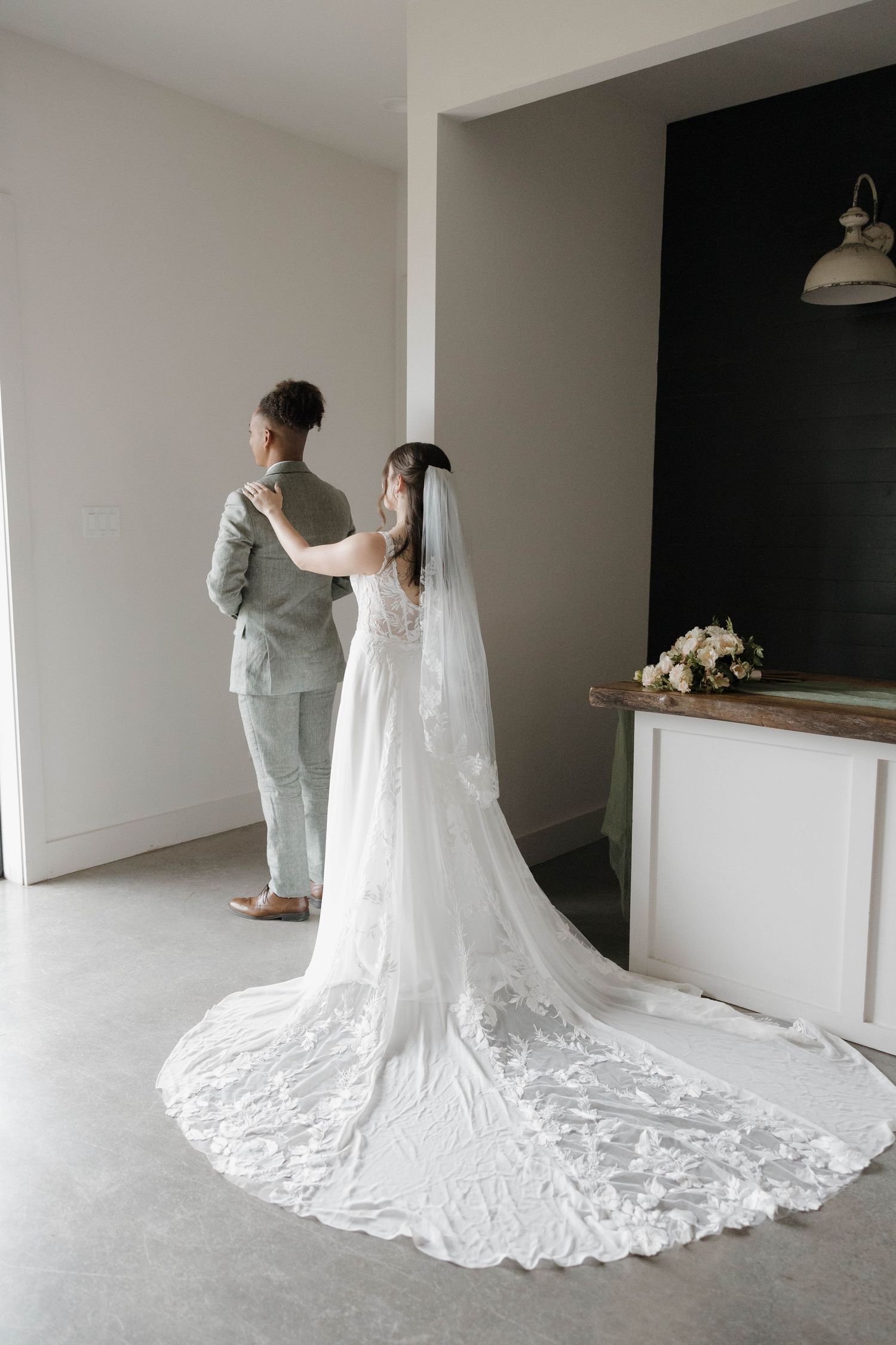 A couple in white wedding attire stand together, showcasing the bride's dramatic lace cathedral veil trailing on the floor.