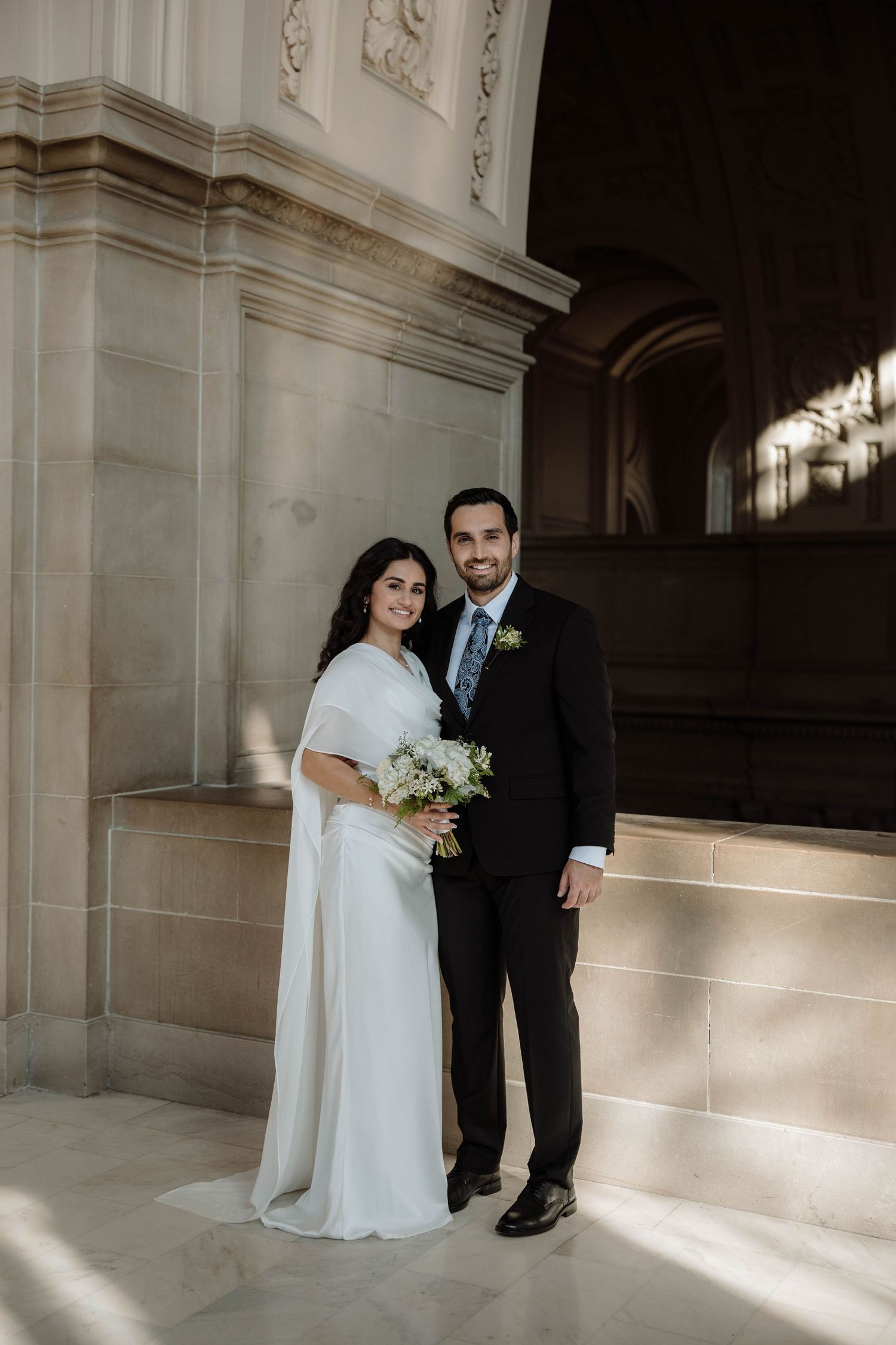 A bride and groom pose together in an elegant indoor setting with ornate white columns and dramatic lighting.