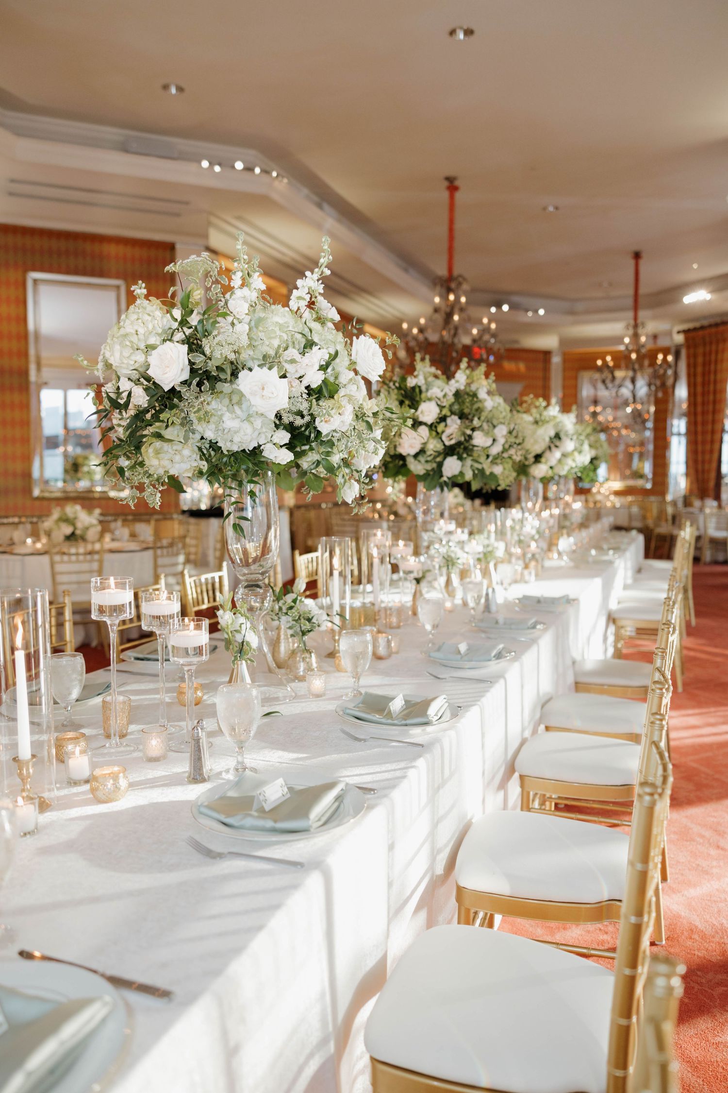 Elegant wedding reception table with tall white floral centerpieces and gold chiavari chairs in a luxurious ballroom setting.