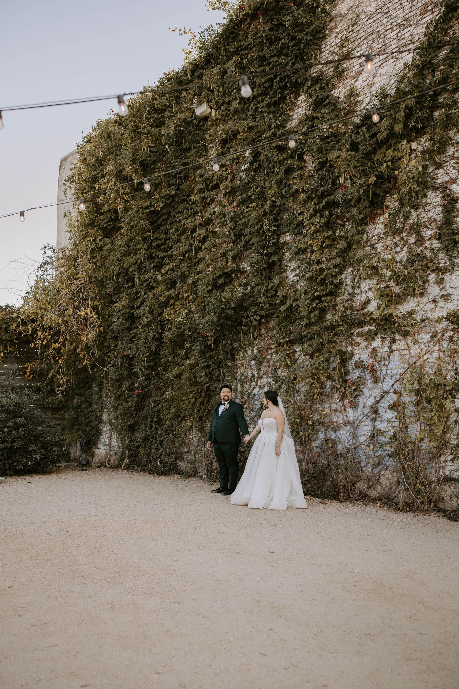Wedding couple stands against tall ivy-covered stone wall on a misty day creating romantic vintage atmosphere.