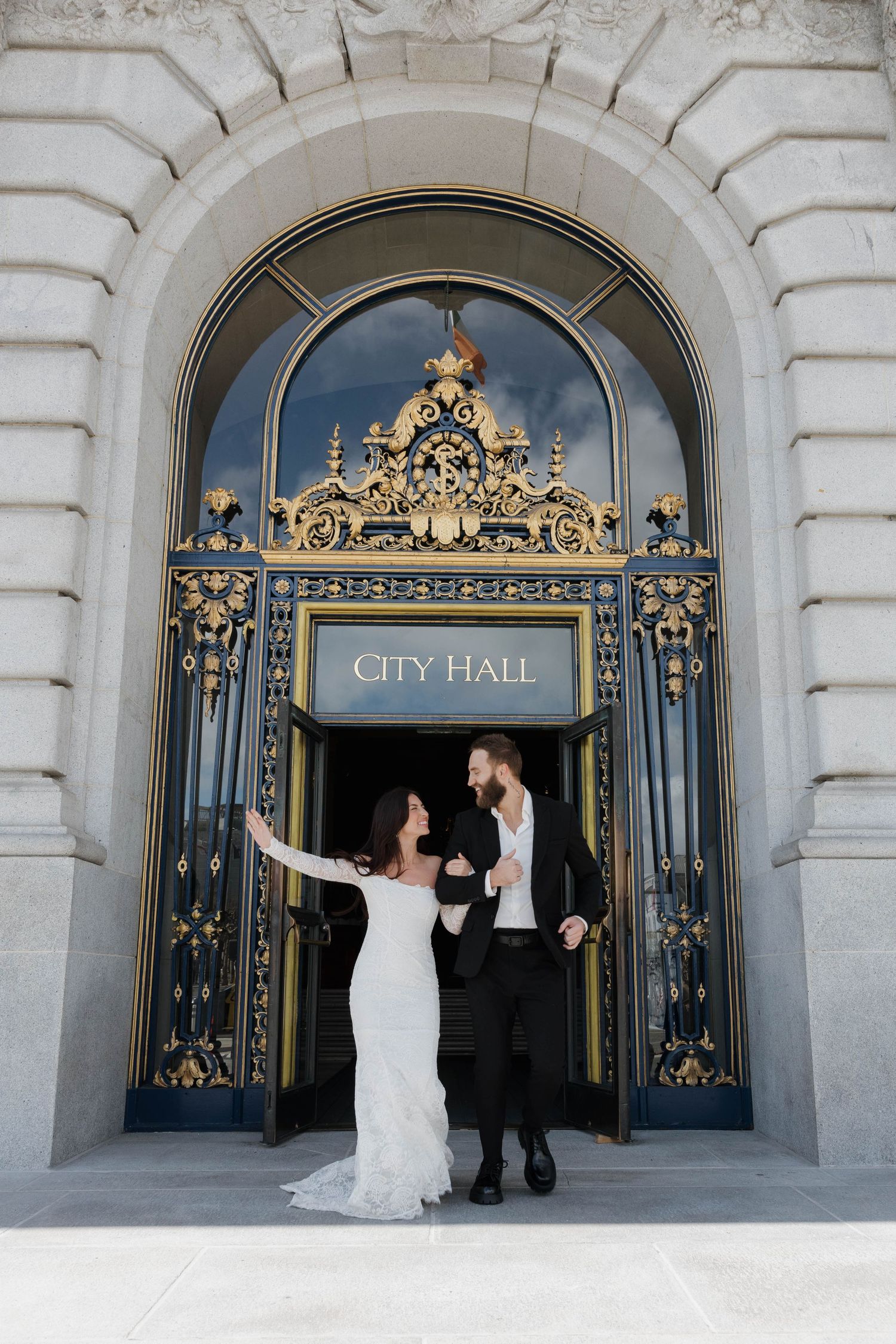 Wedding couple celebrating and raising their arms triumphantly at the ornate City Hall entrance gates.