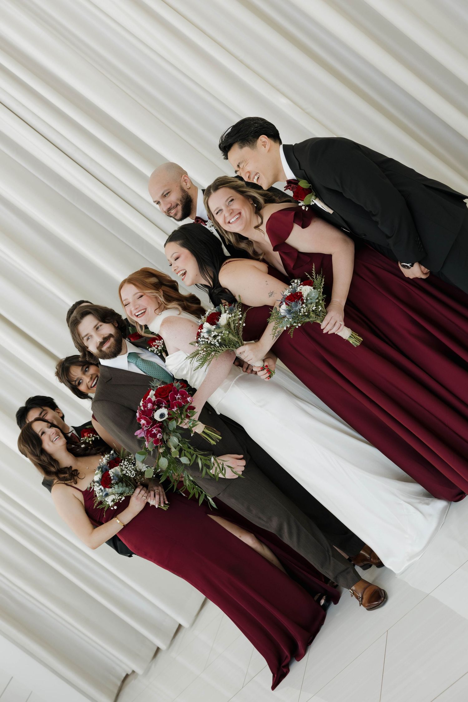 Wedding party in burgundy dresses and dark suits pose together holding floral bouquets against white backdrop.