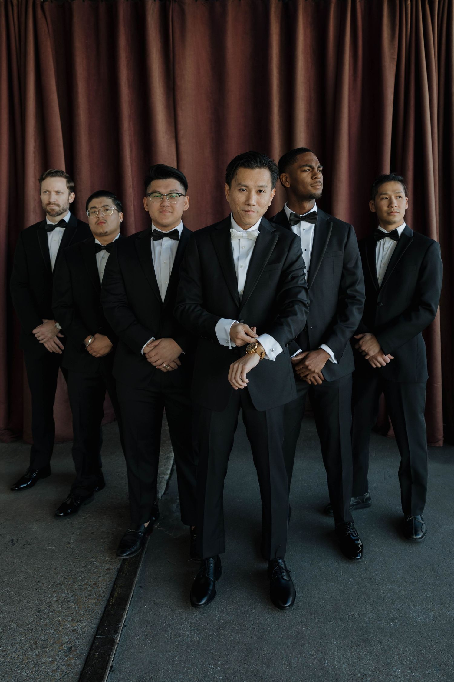 Formal group portrait of groomsmen in black tuxedos against a dark burgundy curtain backdrop.