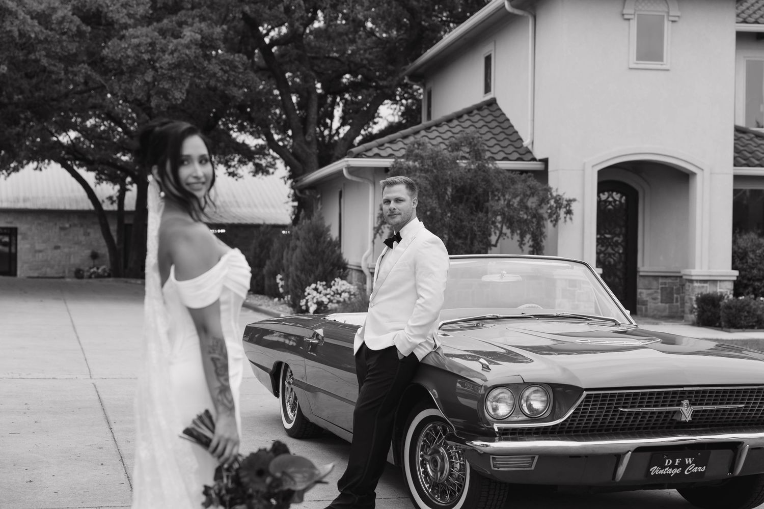 Black and white photo of newlyweds posing with vintage Thunderbird car outside suburban home.