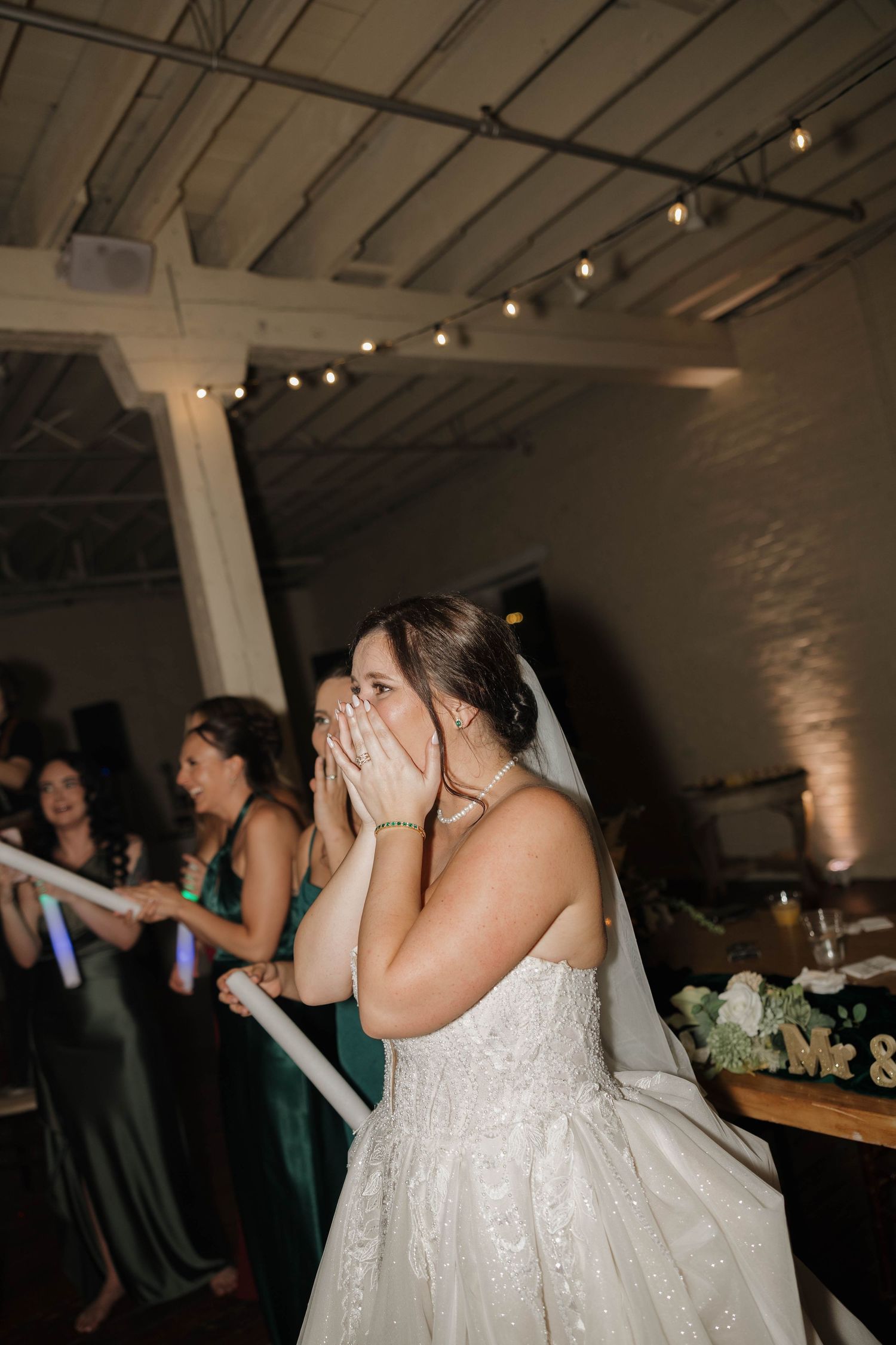 An emotional bride in a white lace gown covers her mouth with her hands while dancing at a reception venue with string lights.