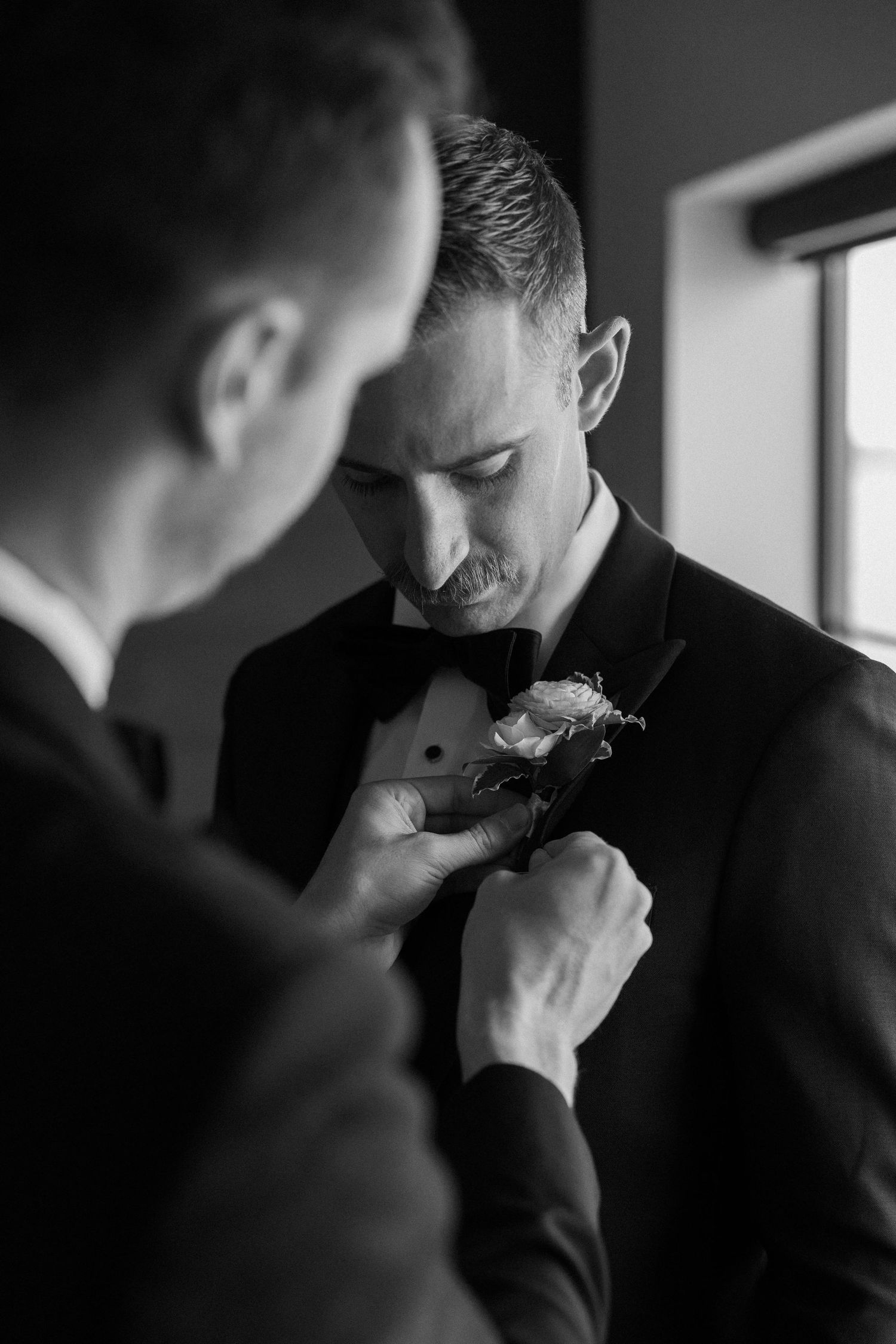 In a black and white photo, someone helps adjust a boutonniere flower on a groom's formal tuxedo lapel.