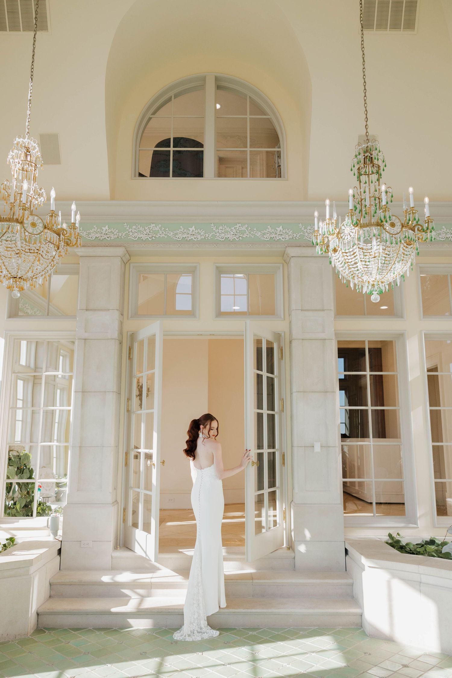 Elegant white wedding gown silhouetted in sunlit doorway of luxury venue with crystal chandeliers and arched windows.