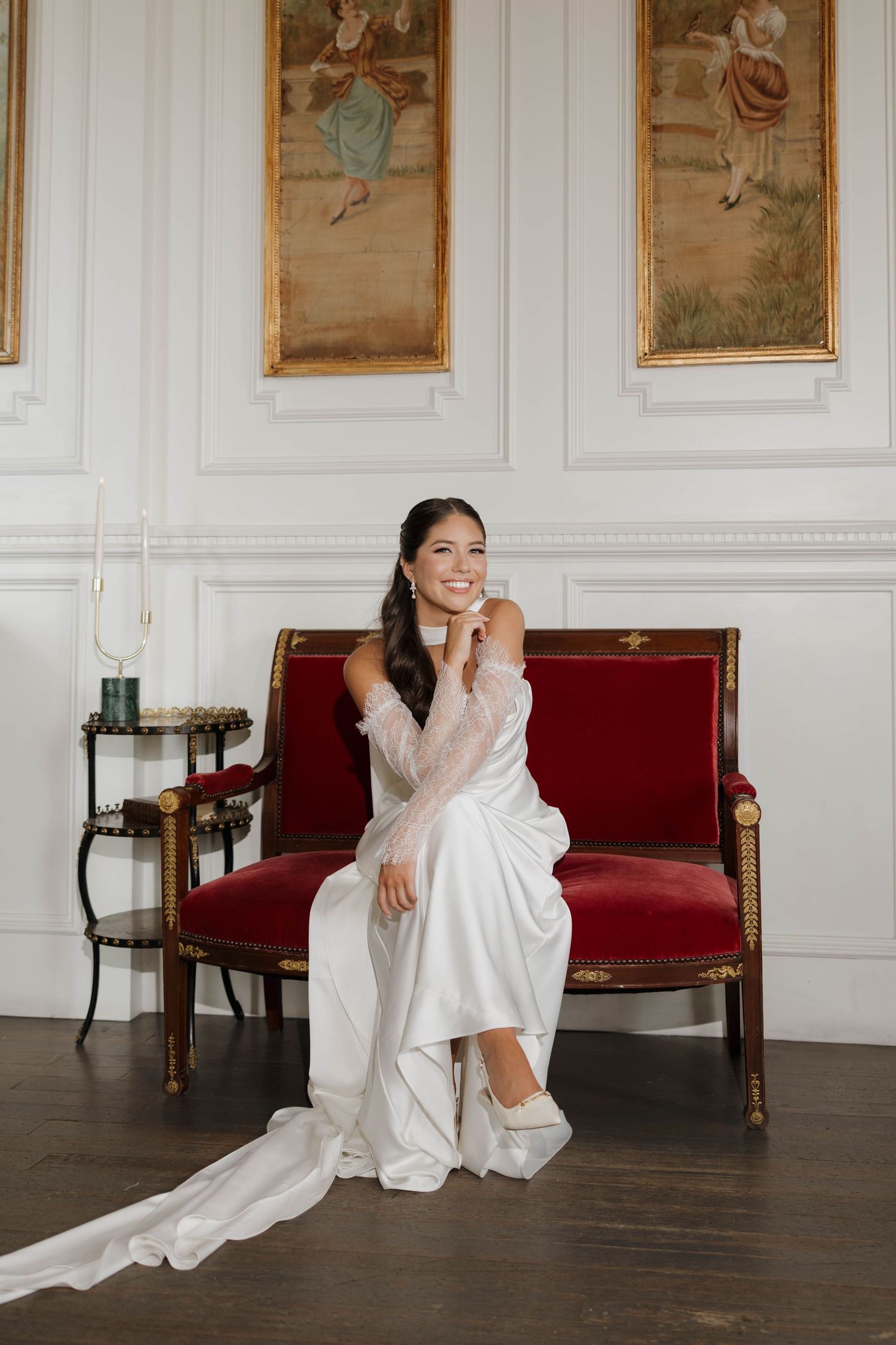 A bride in a flowing white dress sits elegantly on a red velvet bench in an ornate room with framed artwork.