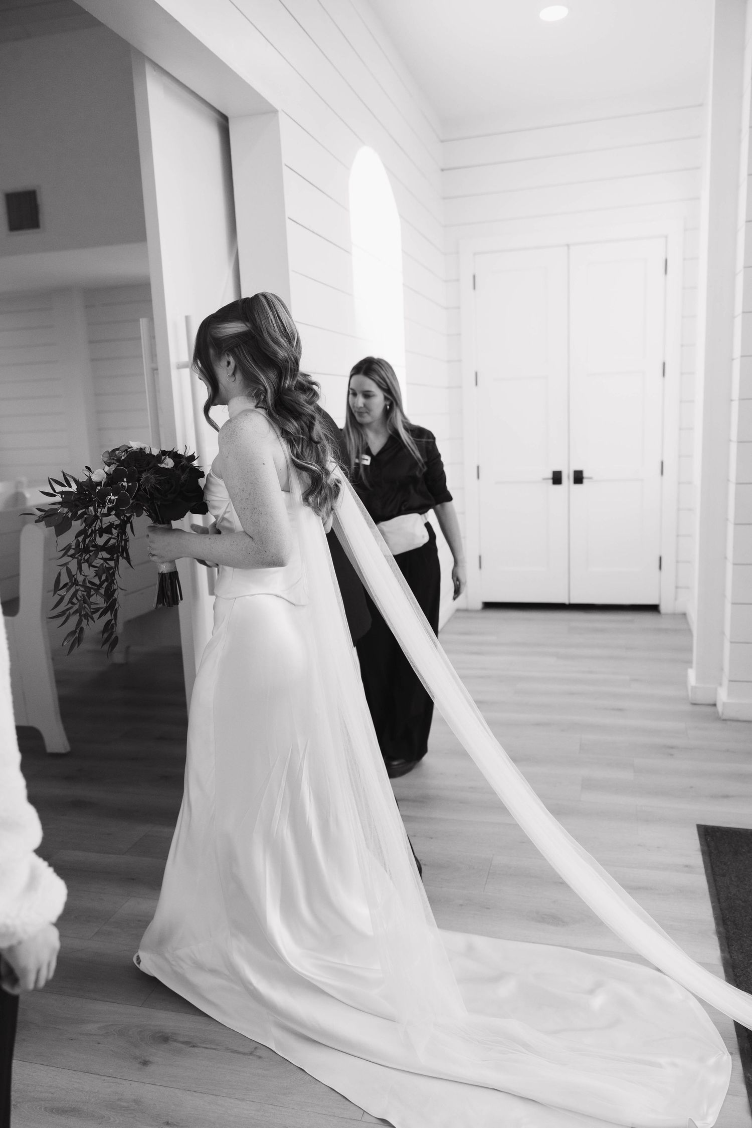 A bride in a flowing white wedding gown with a long train walks down a bright hallway.