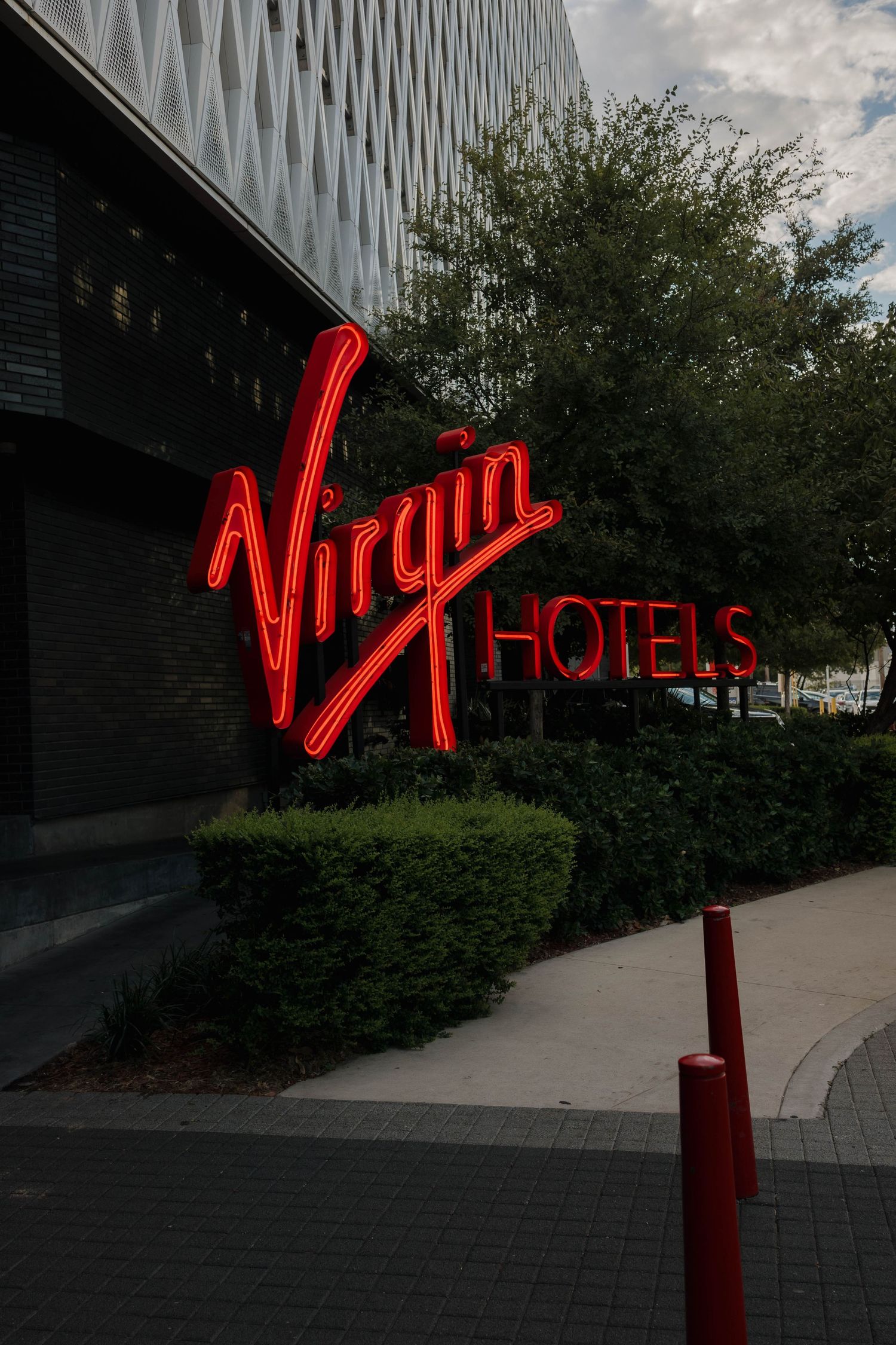 Illuminated red neon Virgin Hotels sign mounted on dark building exterior with landscaping.