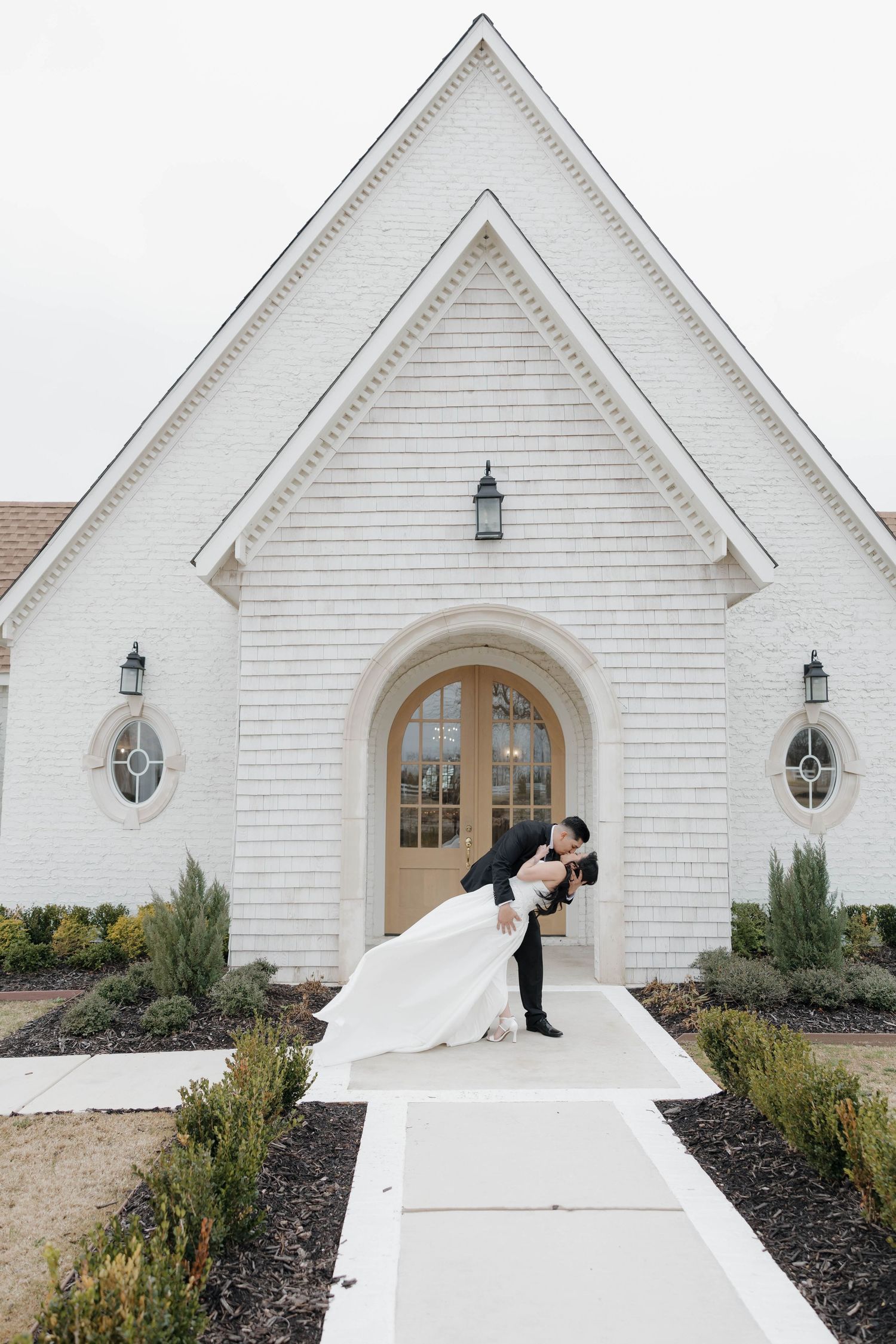 Wedding couple poses dramatically in front of white chapel with arched doorway.