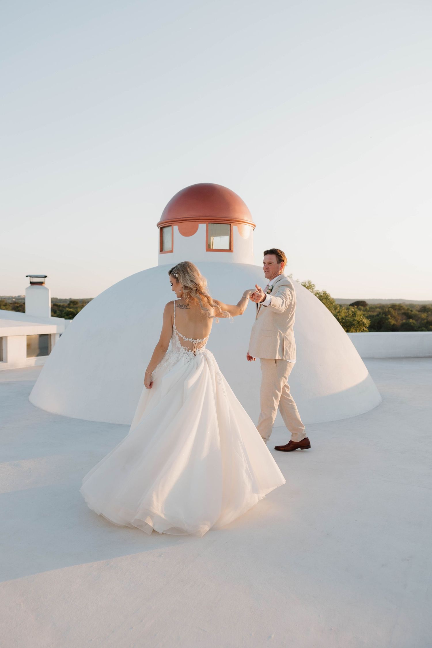 A romantic couple spinning and dancing together on a white domed rooftop against a serene sunset sky.
