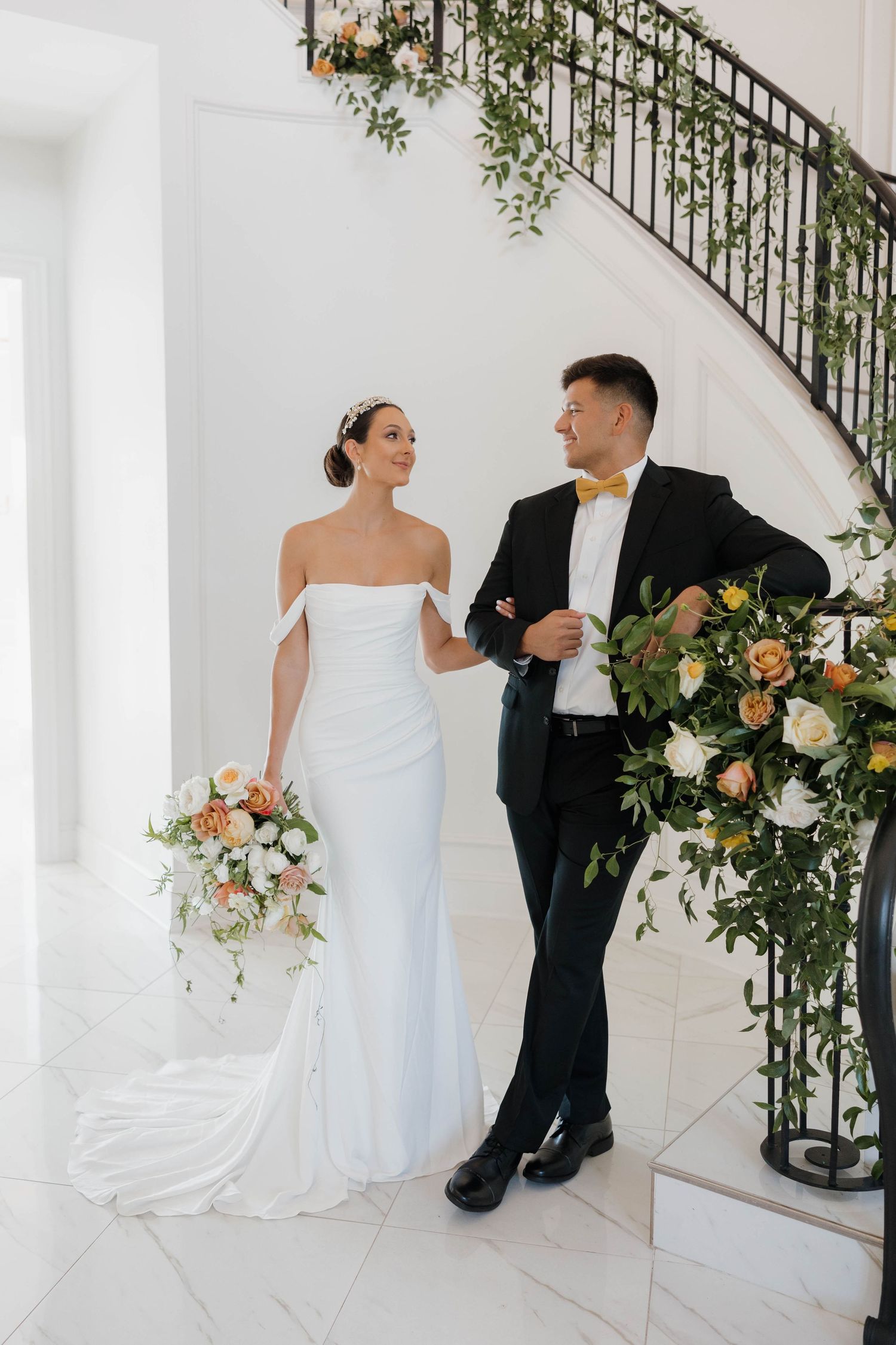 Elegant wedding couple pose on white staircase with floral decorations and greenery.