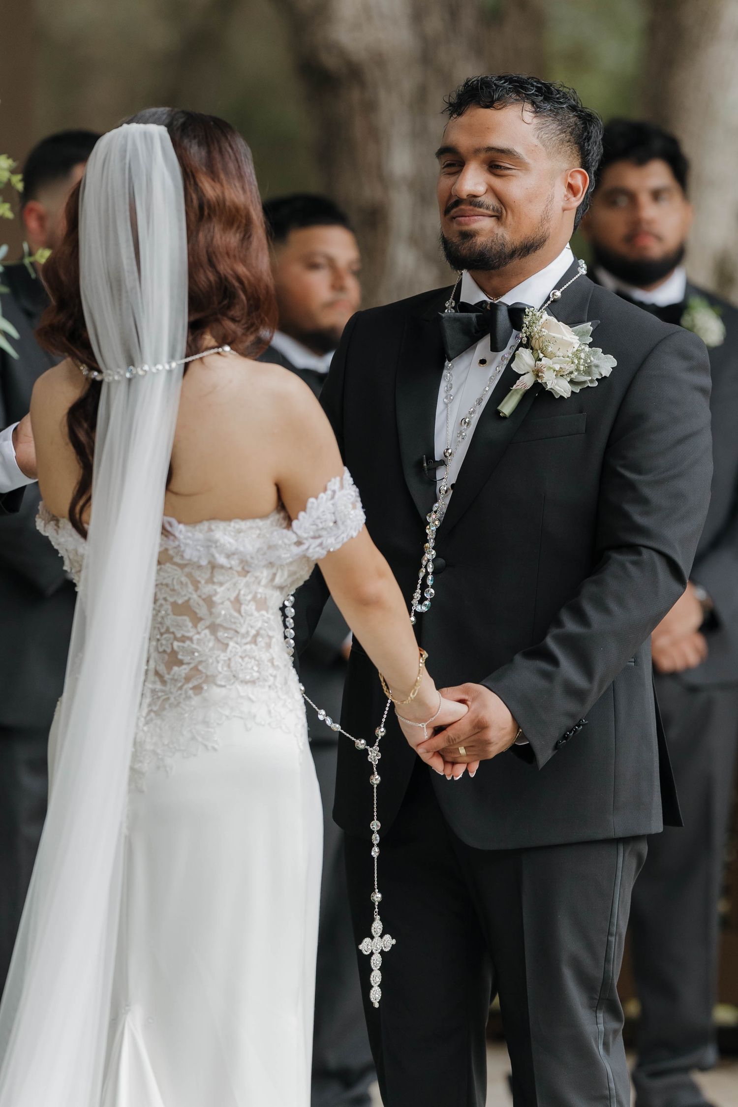 A couple participates in their wedding ceremony dressed in a white gown and black tuxedo with rosary beads hanging between them.