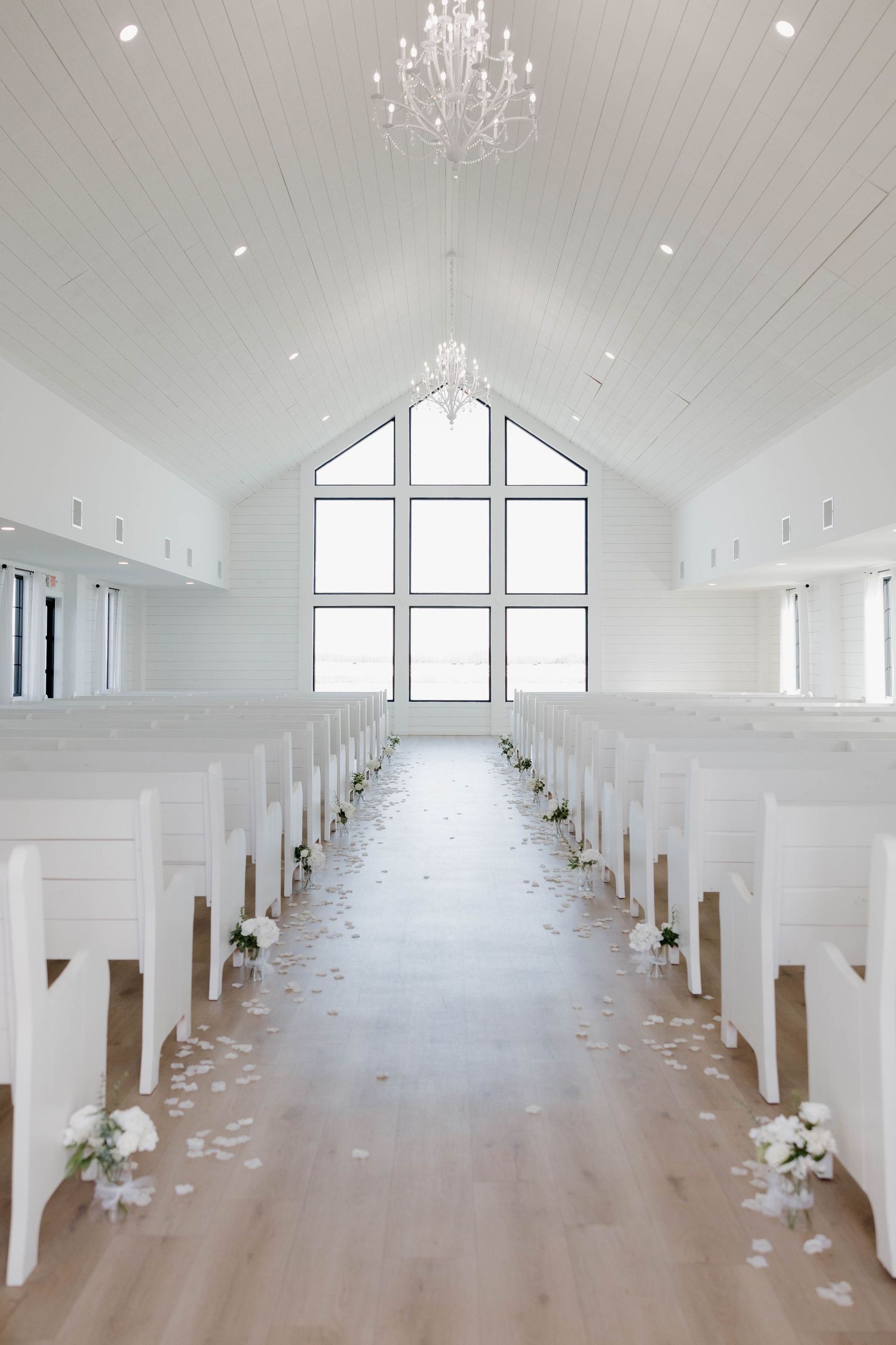 Modern chapel interior featuring vaulted ceilings, crystal chandelier, and white pews lined with flowers.