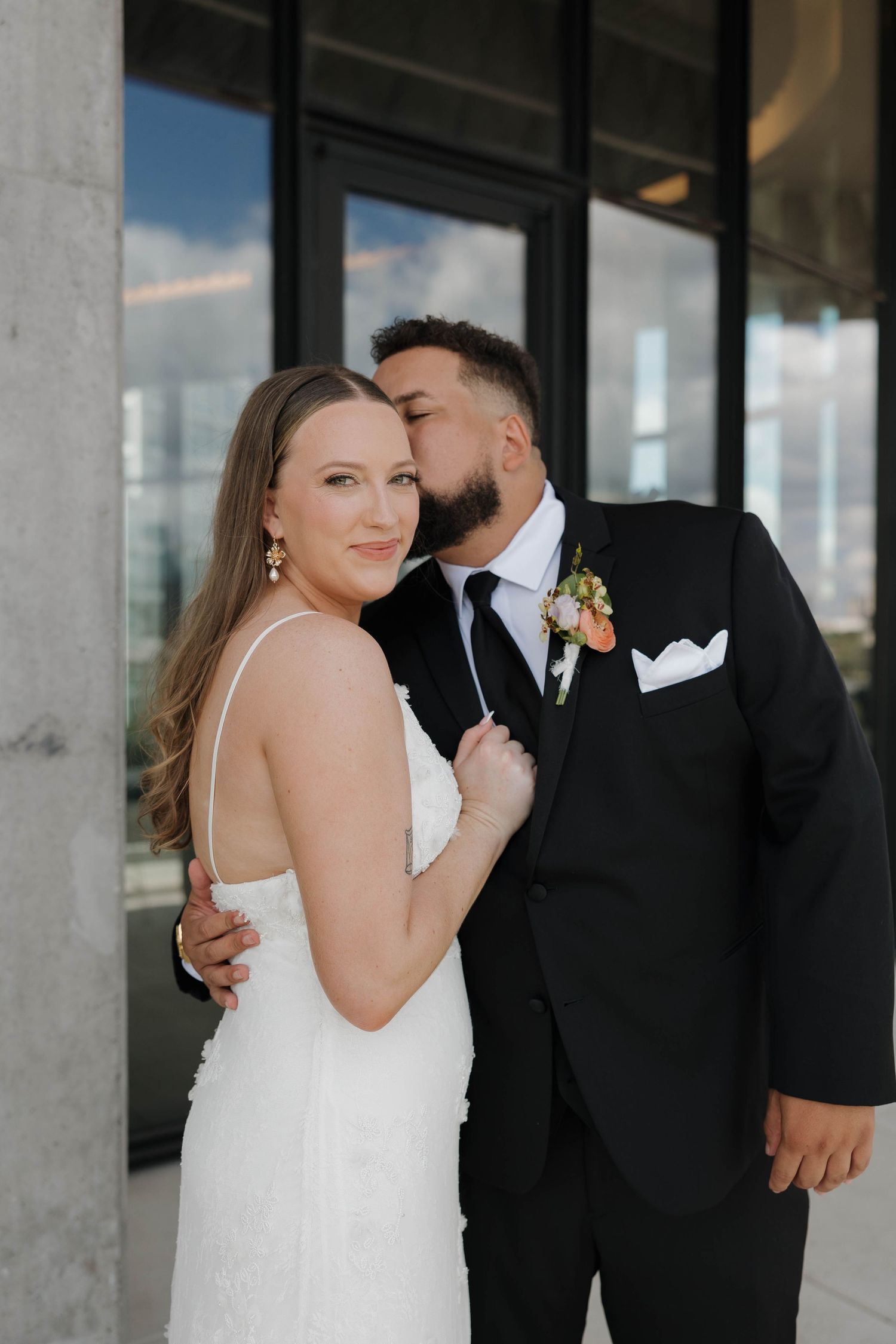 Romantic wedding photo of couple embracing by glass windows with bride in white dress and groom in black suit.