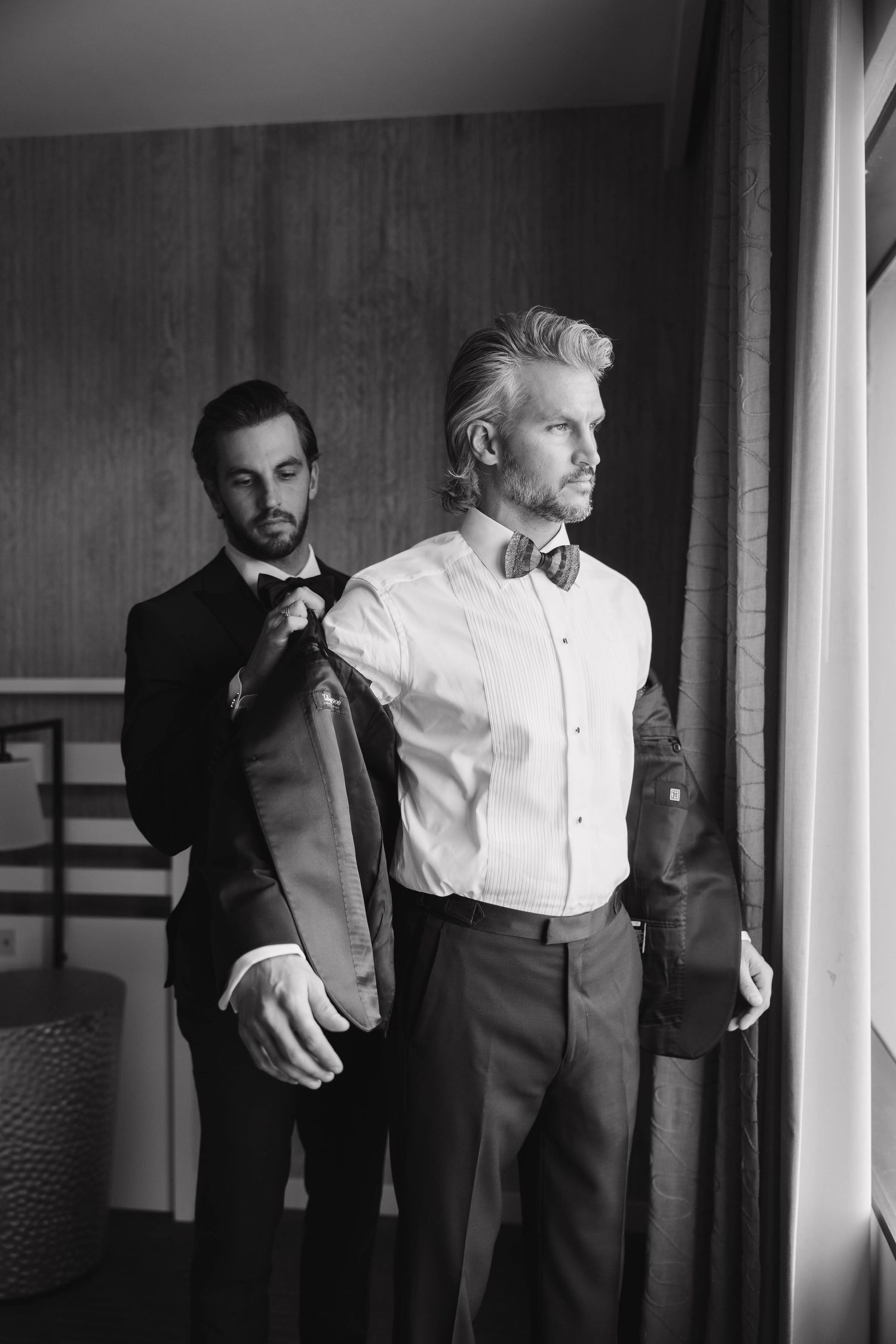 Groomsmen in formal black tuxedos prepare for wedding ceremony in a hallway.