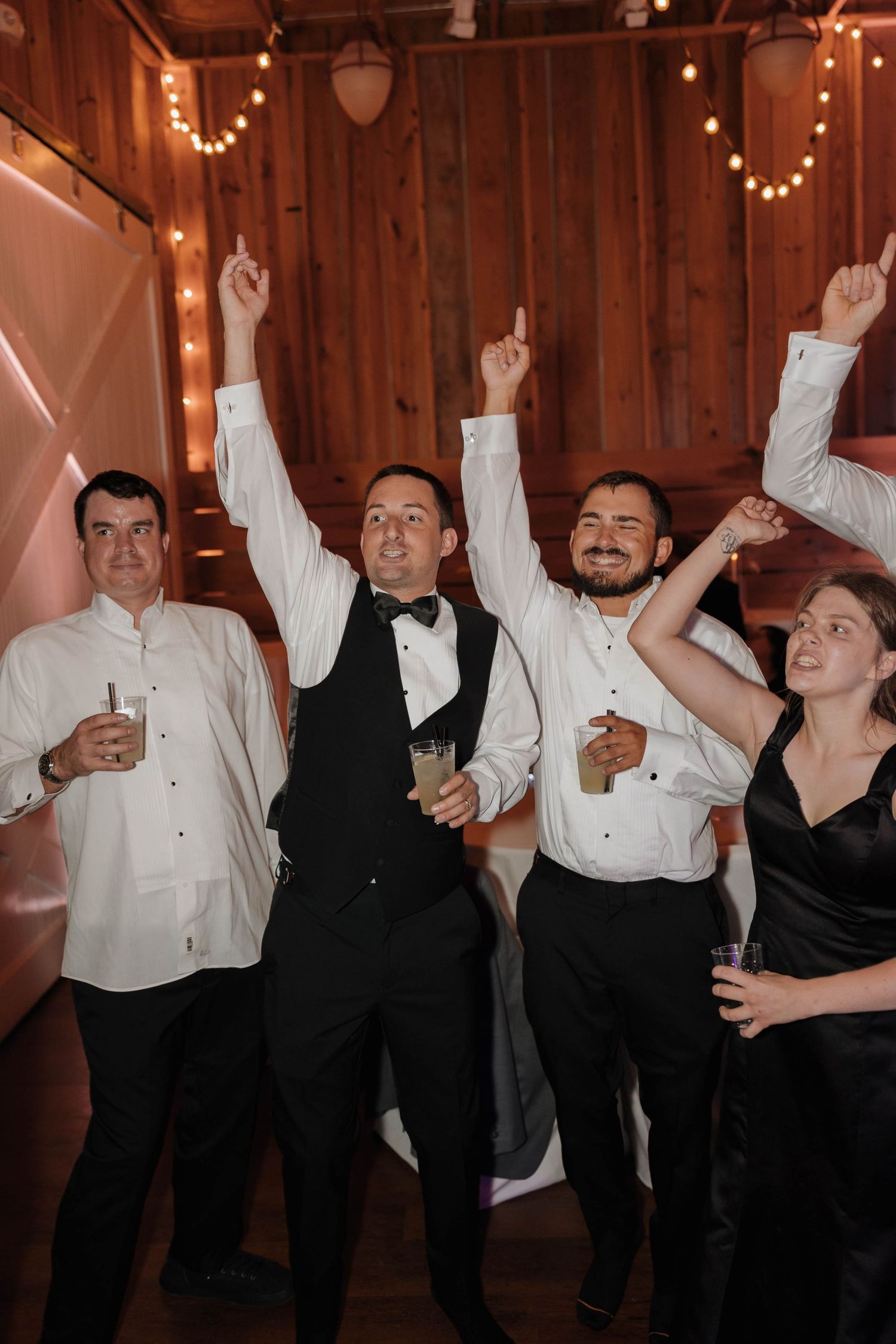 Guests celebrating with raised arms and drinks at a rustic barn wedding reception with string lights in background.