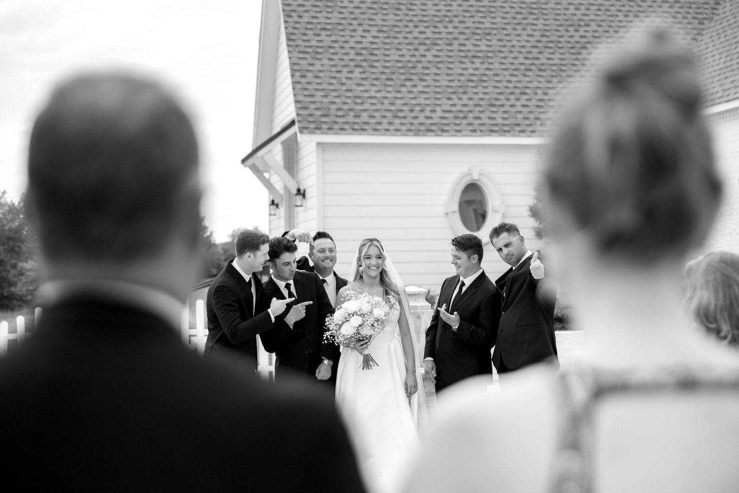 A black and white wedding ceremony photo showing a gathering of formally dressed people outside a white church building.