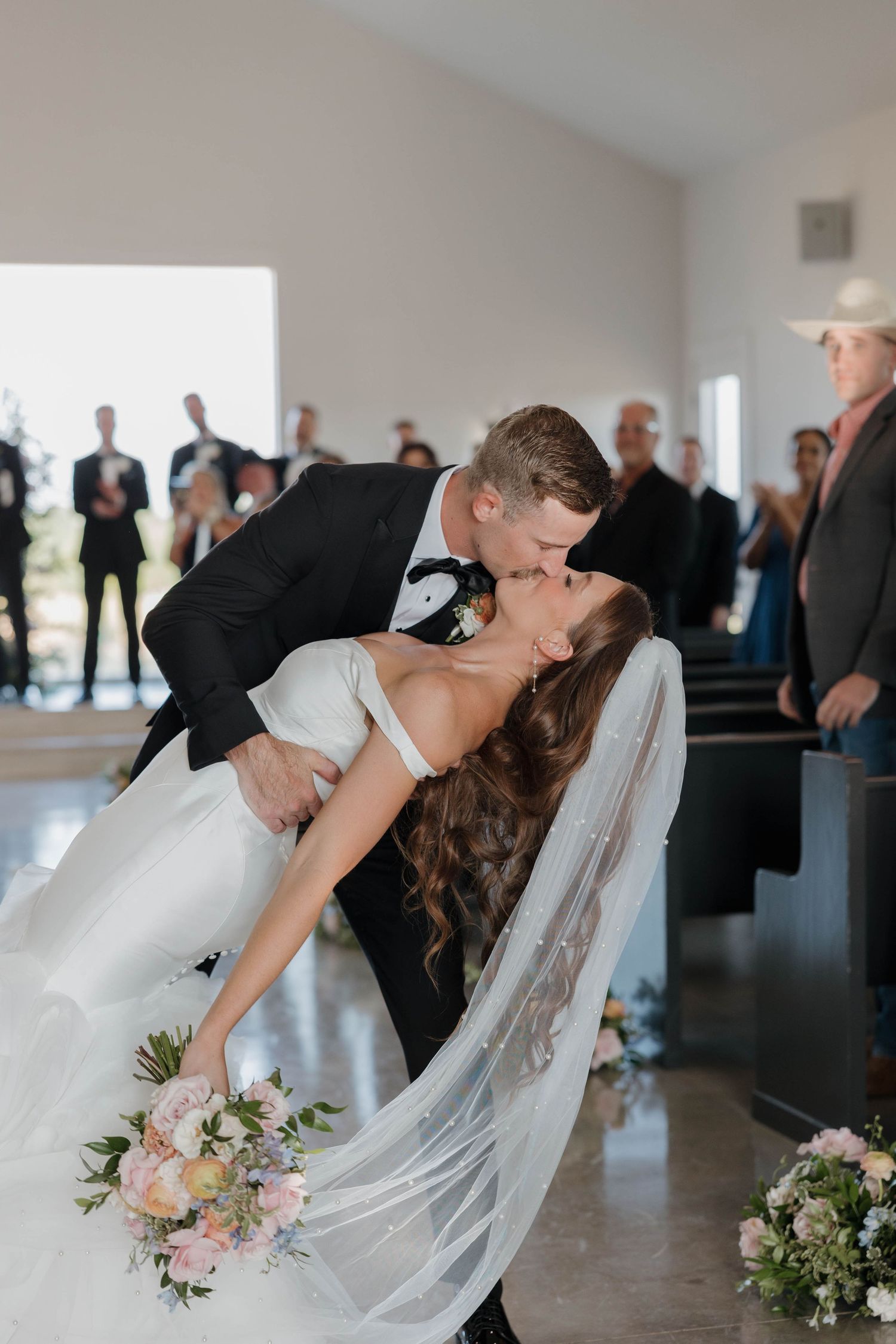 Romantic wedding couple shares passionate kiss in elegant venue with guests watching and flowing wedding veil.