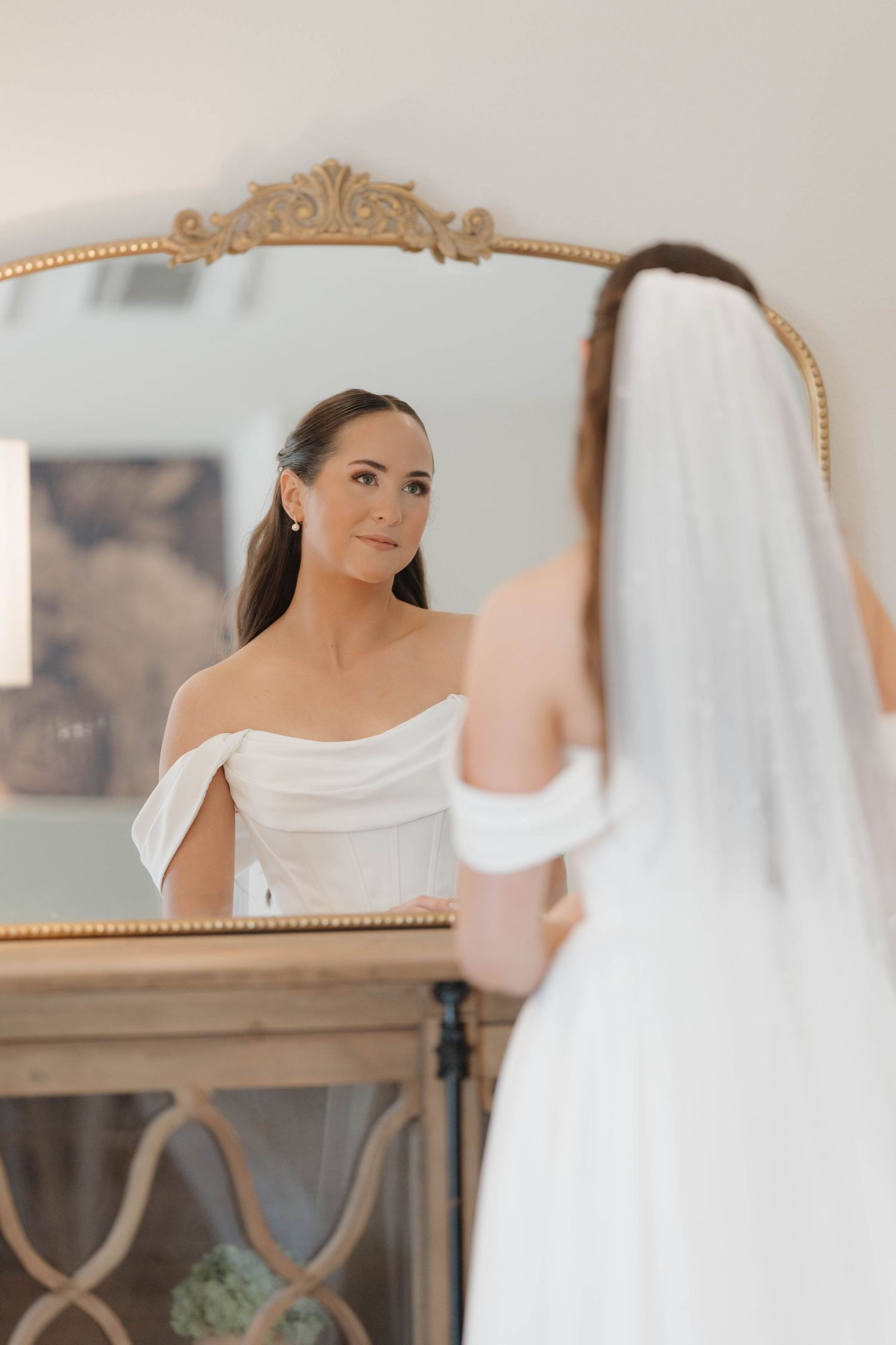 Someone in an elegant off-shoulder white wedding dress looks at their reflection in an ornate gold-framed mirror.