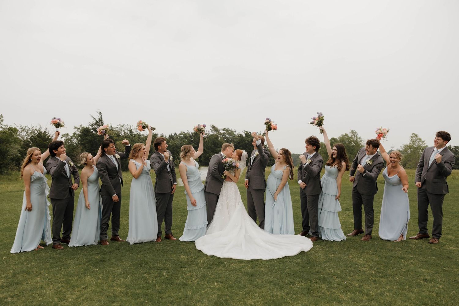 A wedding party stands in a line on a grassy hillside during golden hour, with bridesmaids in light gray dresses.