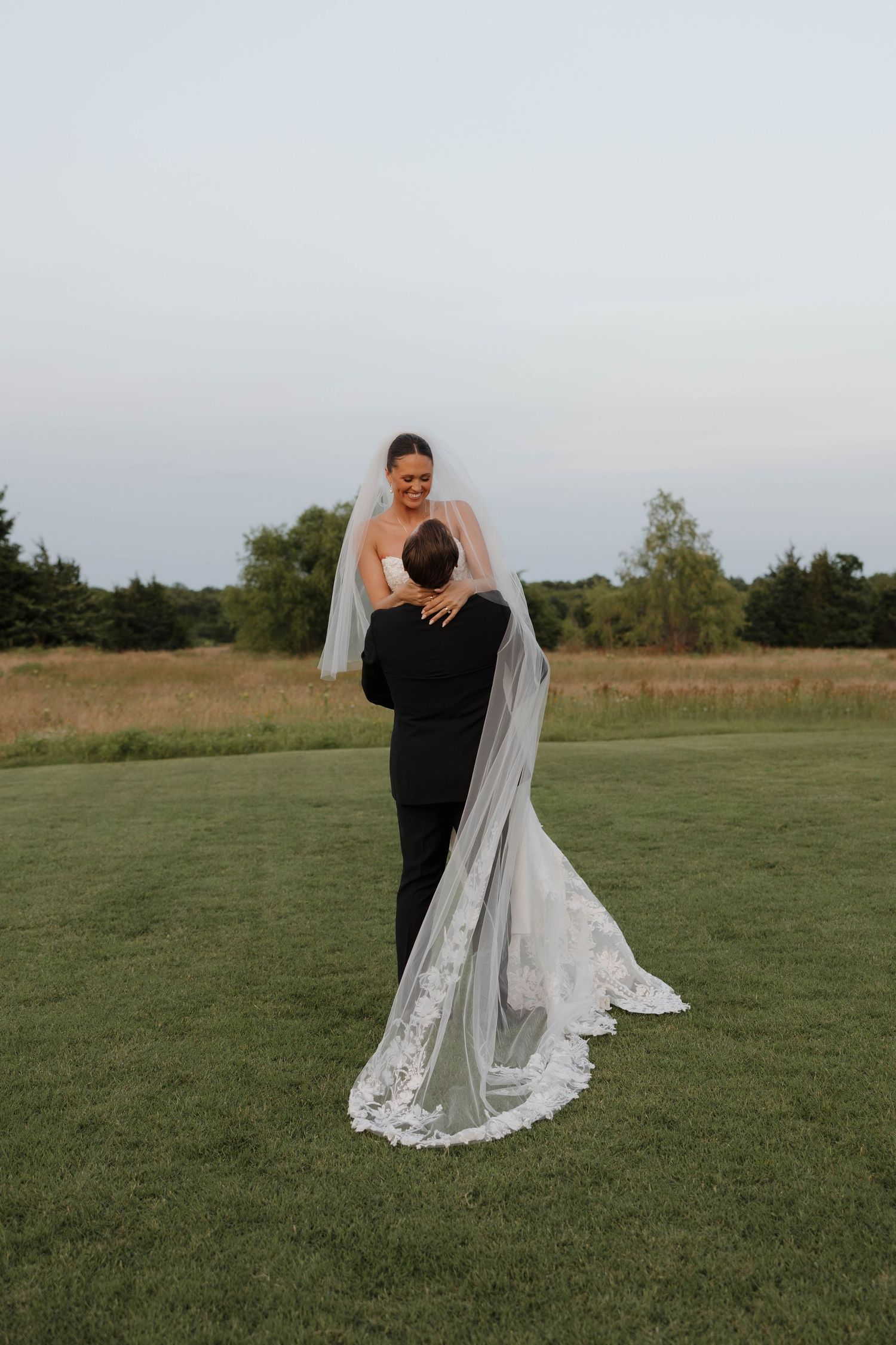 Two people share an intimate moment during a wedding photoshoot outdoors in a grassy field with dramatic lighting.