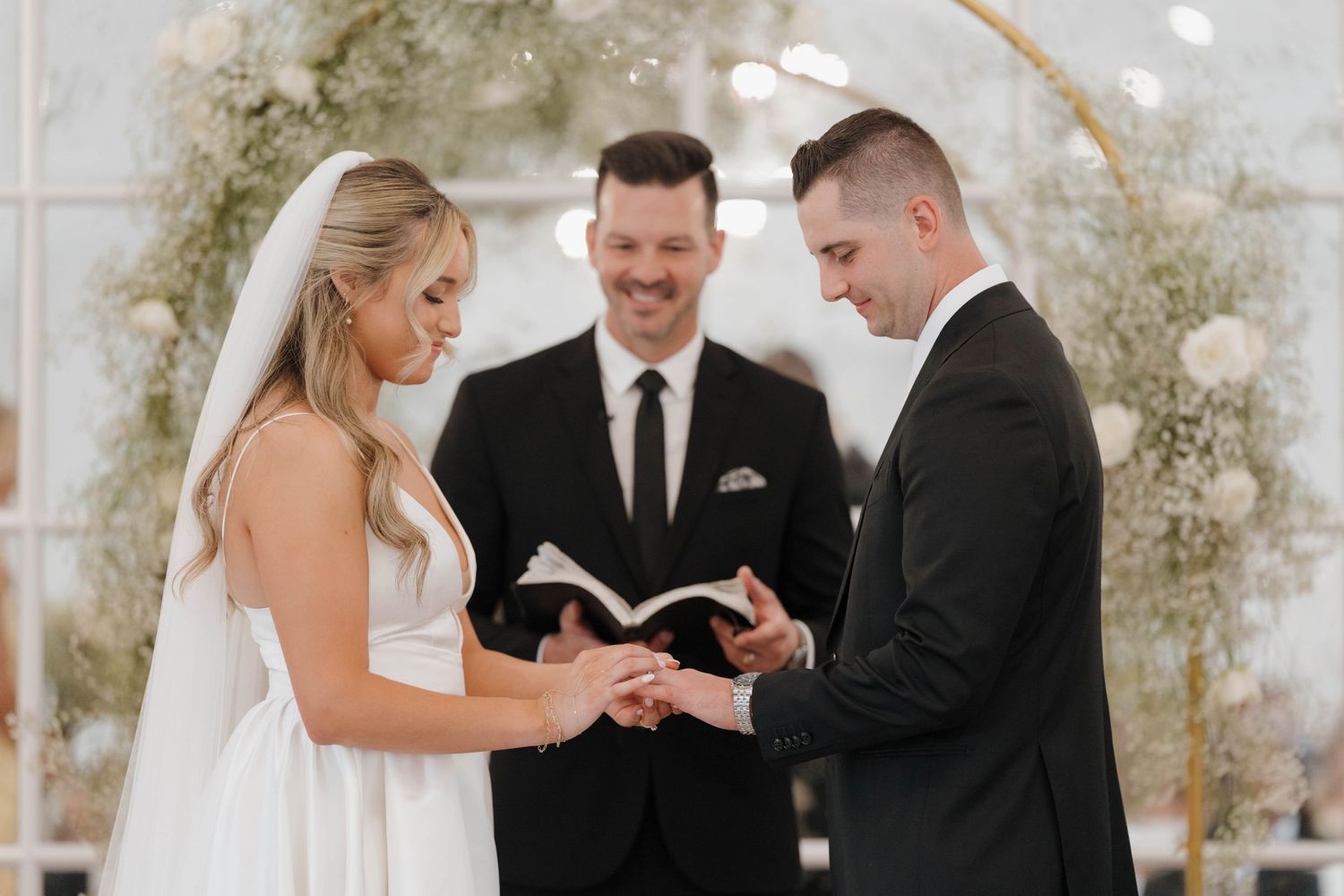 A bride and groom exchange rings during their wedding ceremony beneath a floral arch while the officiant looks on.